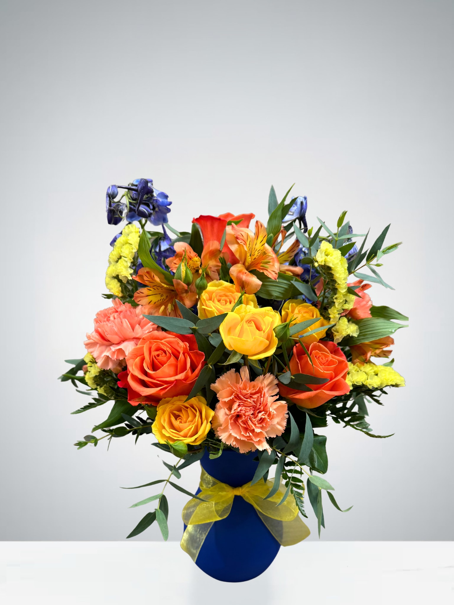 Citrus &amp; Sapphire Blooms - Bring a burst of cheer to any space with this vibrant arrangement! Featuring radiant orange roses, golden yellow spray roses, peach carnations, and bright alstroemeria accented with touches of deep blue delphinium and sunny yellow statice, this bouquet is designed to brighten the day. Arranged in a striking cobalt blue vase with a cheerful yellow bow, it’s a perfect gift for celebrations, thank-yous, or simply to spread joy.