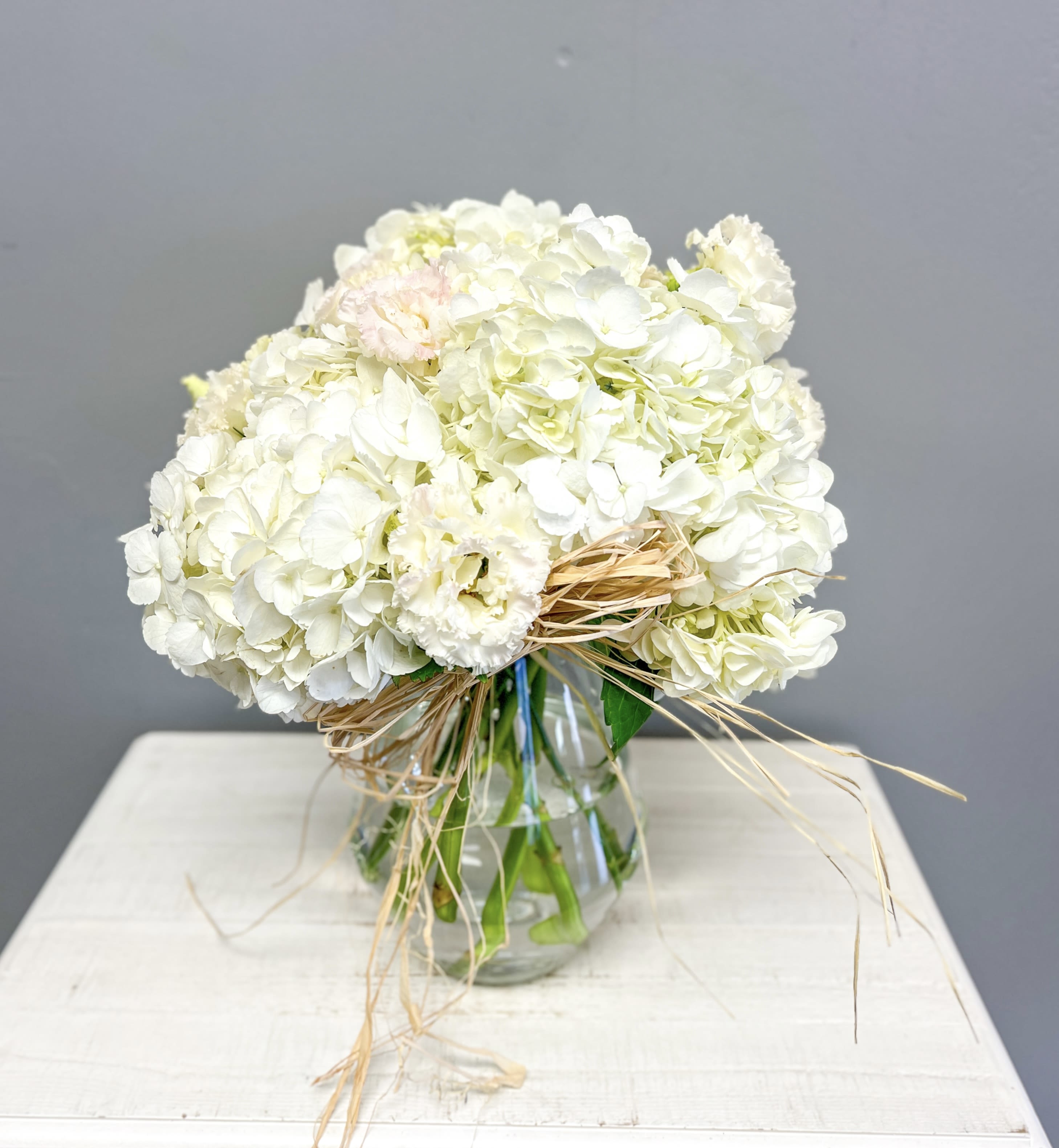 Whispers of Grace Whispers of Grace - Let simplicity speak volumes with our Whispers of Grace arrangement—a lush medley of white hydrangeas and soft blush-toned lisianthus, hand-tied with natural raffia for a rustic, heartfelt finish. Designed in a clear glass vase, this timeless bouquet is perfect for sympathy, weddings, baptisms, or simply sharing peace and love.  Crafted with care by Anointed Flowers & Gifts in Jackson, TN.