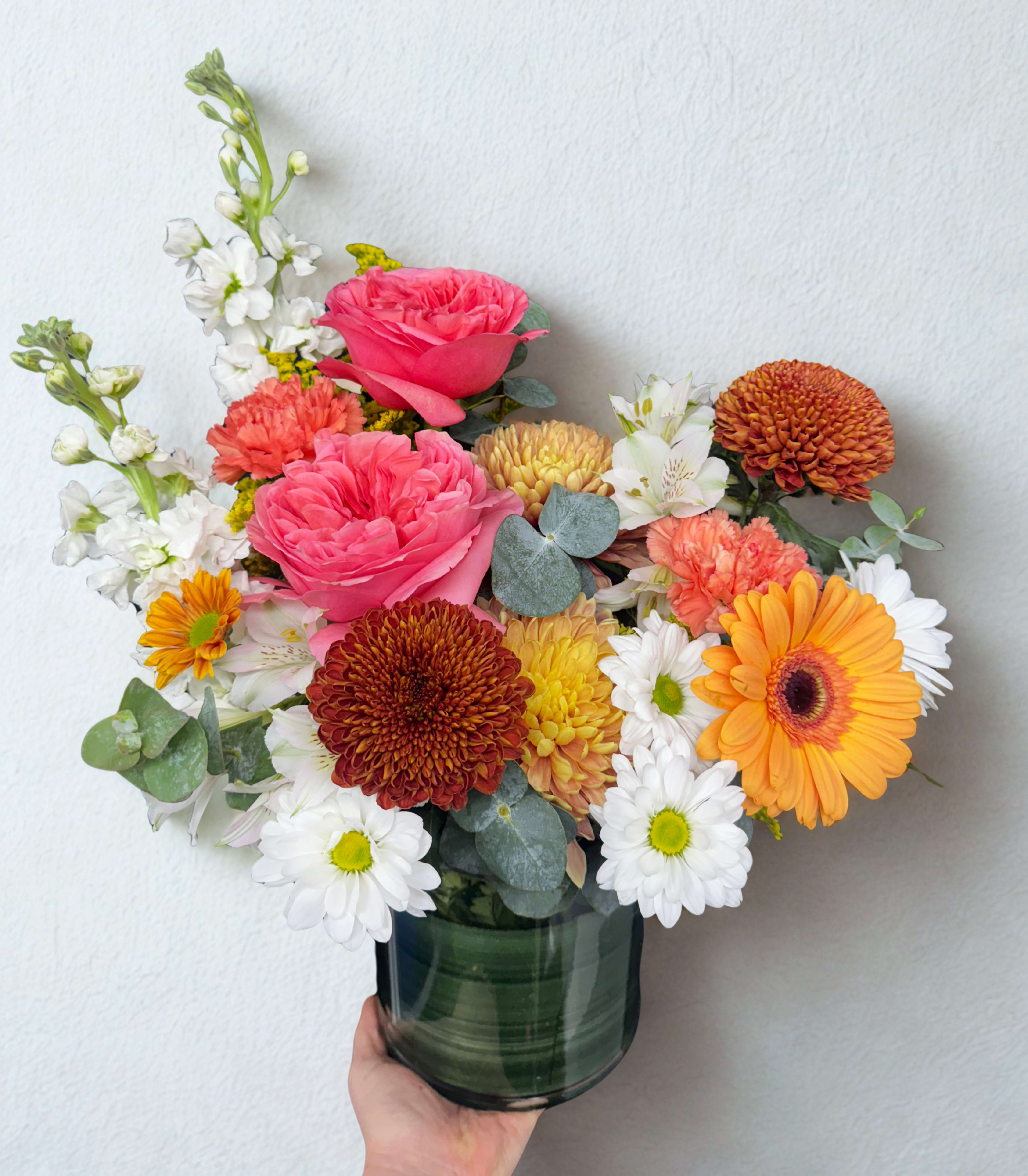 Sunset Bloom Sunset Bloom - A radiant mix that feels like golden hour in a vase. featuring garden roses, gerbera daisies, chrysanthemums, carnations, stock, alstroemeria, daisies, and eucalyptus. Warm tones of peach, coral, orange, and white blend for a bright, uplifting design that fits any space or occasion. Note: Garden rose may be replaced with a premium bloom of equal value based on seasonal availability.