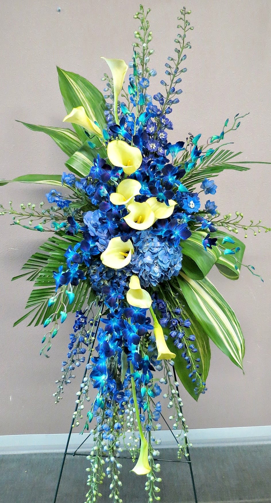 Ocean Blue - This is one of our beautiful easel arrangements. Designed with delphiniums, hydrangeas, dendrobium orchids, calla lilies, aspidistras, and palms. Dimensions: 60&quot;x25&quot;