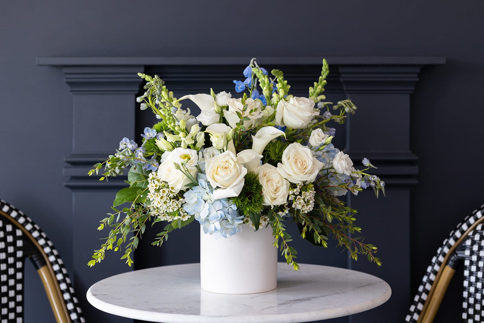 By the Sea Collection - Signature Size  - Nothing restores a sense of joy and rejuvenation like spending some time oceanside. Our By the Sea Collection is a tribute to a seascape filled with shades of blue and white. Each arrangement is overflowing with hydrangea, mini Callas lilies, delphinium, Snapdragons, Dianthus, Lisianthus, Snapdragons and fresh Eucalyptus.   Our J. Morris Signature designs are perfect for an anniversary, birthday, as a thank you, or as a larger centerpiece for a kitchen island or table. They are low enough to see over but are still full, grand and filled with blooms. Featured in a matte white 6” ceramic vase.   Dimensions: J. Morris Signature arrangements are 15” high and 12 inches wide.  