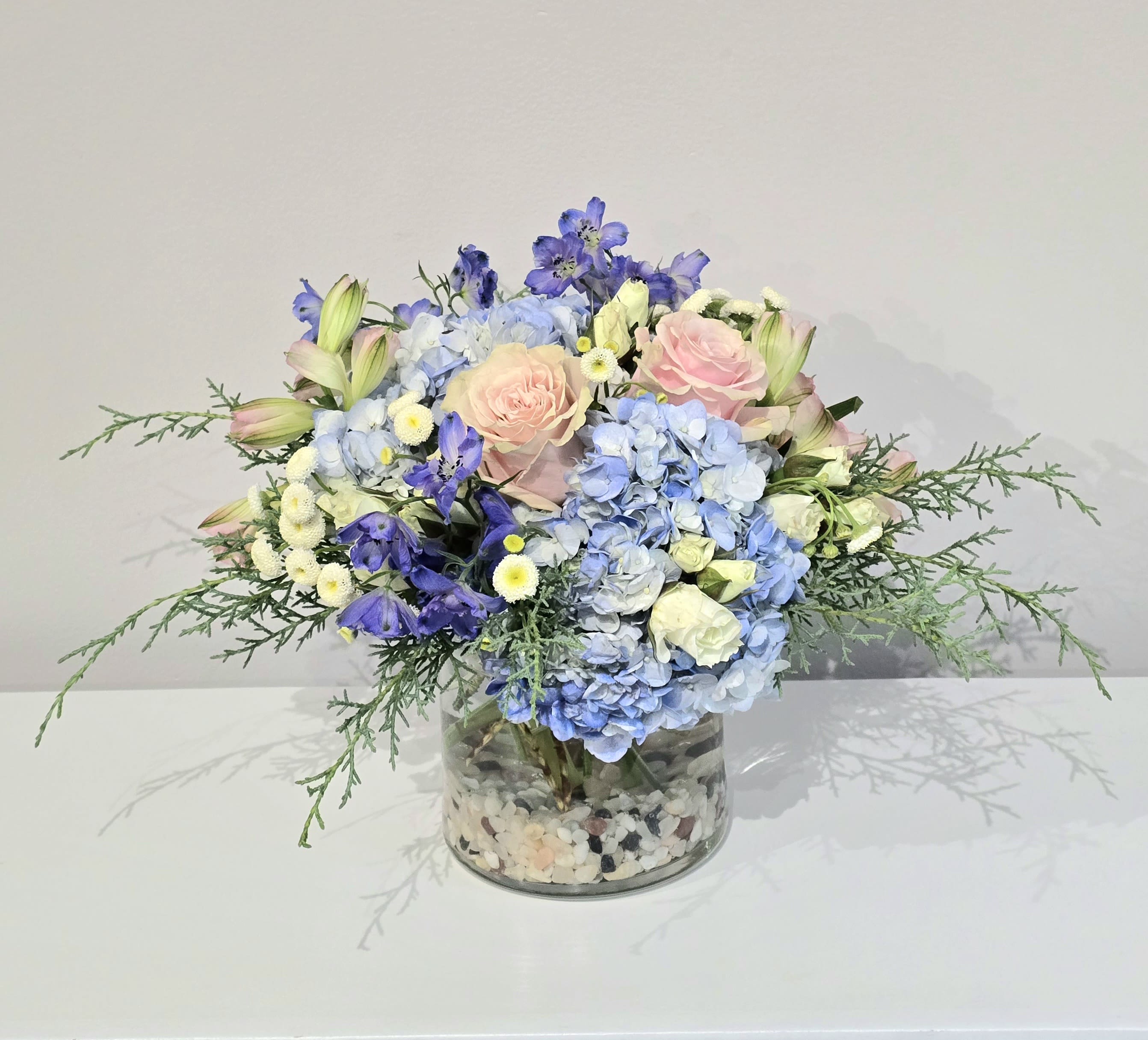 The Rivers Edge - A fresh blue, white &amp; light pink arrangement that features seasonal blooms like, blue delphinium &amp; hydrangeas, pink roses, white spray roses, delicate white chamomile, pink alstroemeria, &amp; fragrant cypress evergreens set inside a clear glass cylinder vase accented with river rock pebbles. Arranges low &amp; compact. An elegant bouquet appropriate for all occasions. Makes a lovely centerpiece display. 