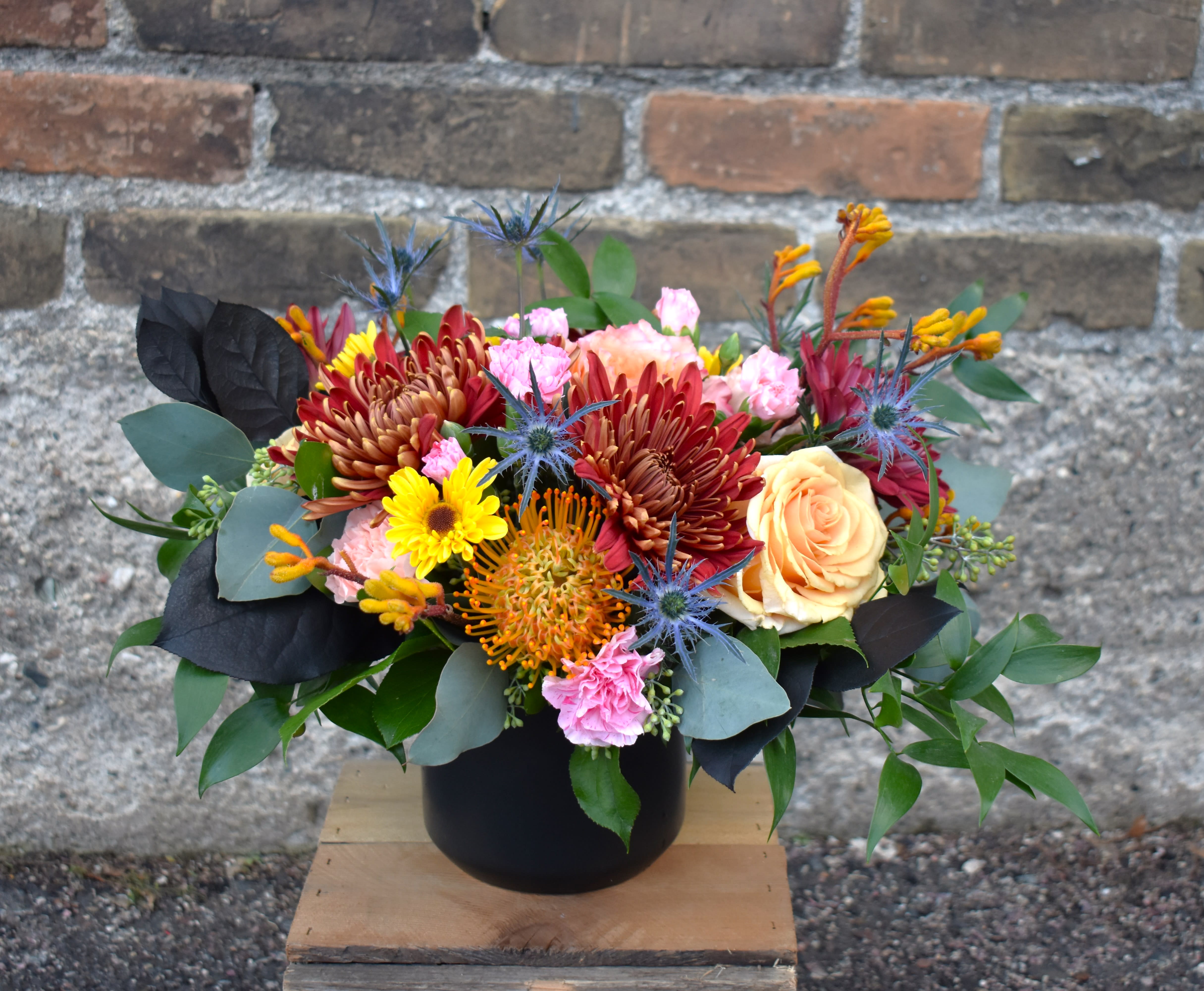 Ember Ember - Brilliant hues and varied textures form this rich bouquet! peach roses, bronze cremone mums, yellow viking pomps, blue thislte, pink miniature carnations, orange pincushion protea, gold kangaroo paw, and lush greens are designed in a black ceramic vase and accented with black-tinted salal...a luxurious way to celebrate the season!