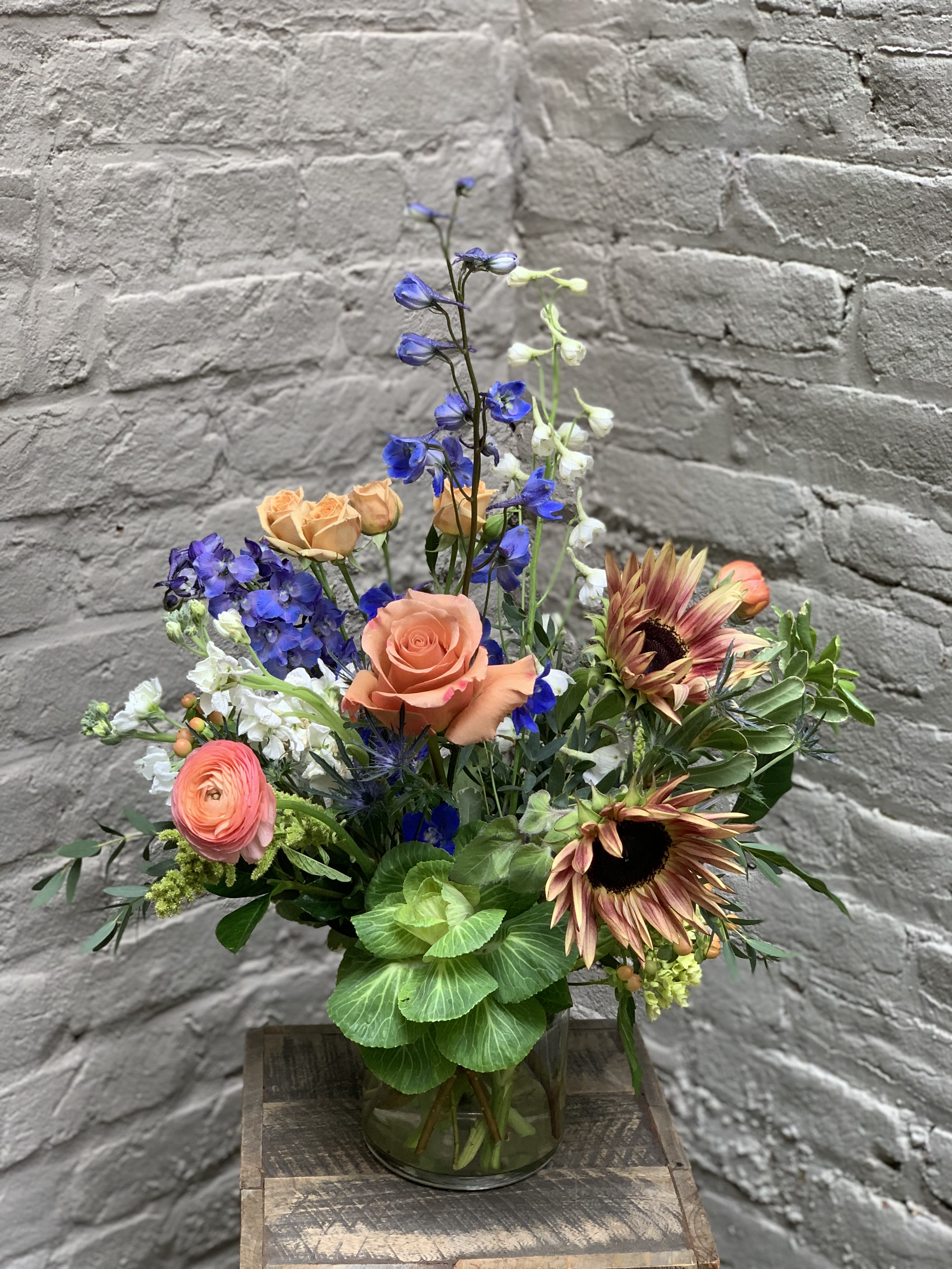 Afterglow Afterglow - A lush bouquet of soft flowering blooms designed to grace your tabletop or tailgate spread. This arrangement features a colorful, seasonal palette of Moab roses and plum sunflowers accented by deep blue delphinium all set off by a mix of textured foliage in a 5” x 5” glass cylinder. Arrangement measures approximately 12" x 14".
