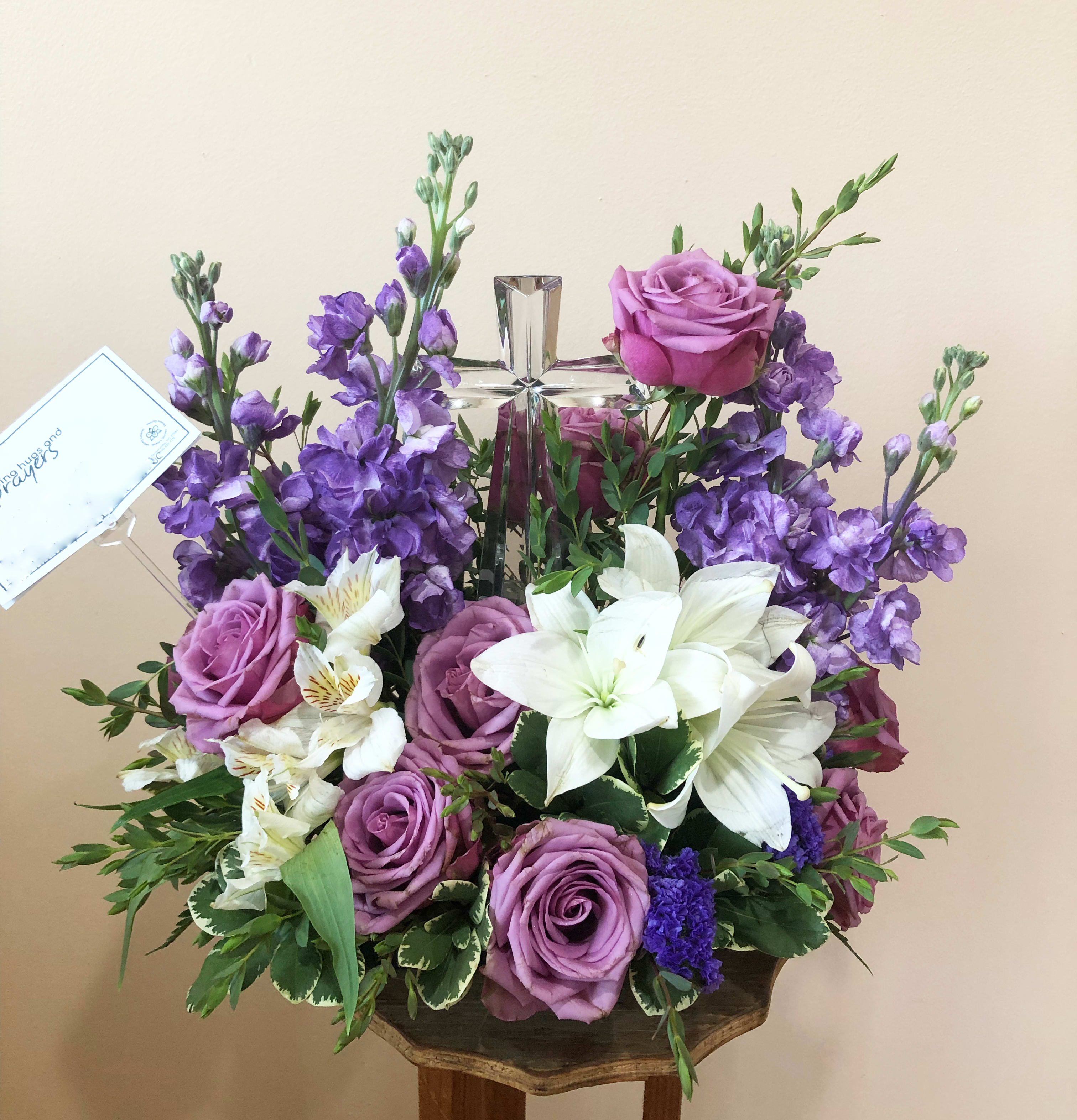 Poised with Love Bouquet - Soft and serene, this artistic array of cream and lavender blooms around a radiant crystal cross keepsake is a heartfelt expression of love. This beautiful piece includes crème roses, lavender larkspur, lavender montecasino asters, white cushion spray chrysanthemums, lavender sinuata statice, lemon leaf and dusty miller. Delivered with Teleflora's Small Crystal Cross.