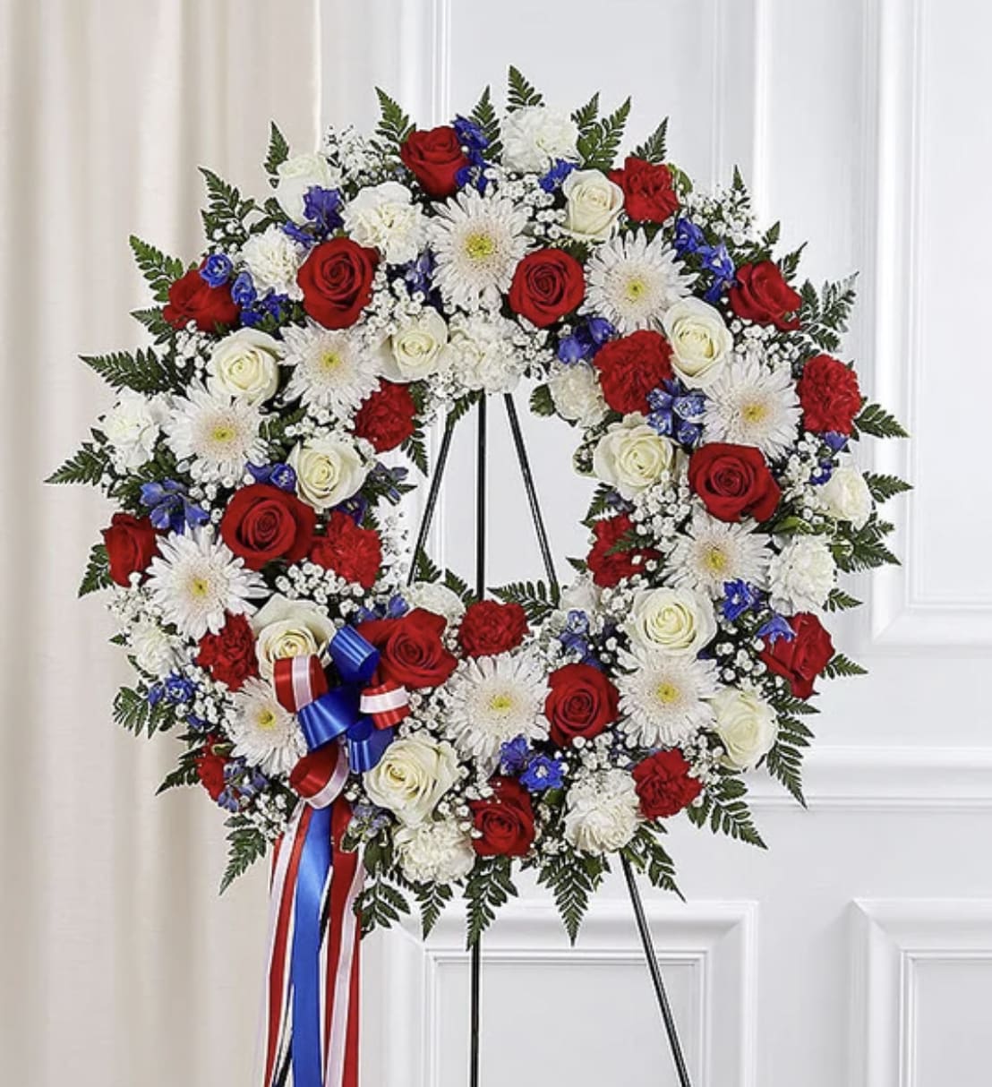 Serene Blessings Standing Wreath- Red, White &amp; Blue - They served their country with honor and pride, so it’s only fitting to honor them with a beautiful symbol of eternal life. Our patriotic standing wreath arrangement is meticulously crafted by our expert florists to honor a brave veteran who has passed away. Filled with lush blooms in red, white and blue, it’s a fitting final tribute for the funeral services.
