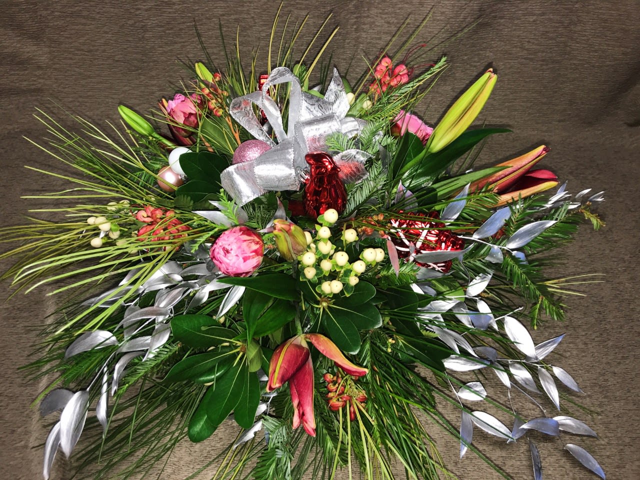 Christmas Ornament Centerpiece Large with Silver Foliage - This semi round arrangement of holiday greenery, berries, premium flowers and lilies will be long lasting so enjoy it as we lead into Christmas week or into the New YEAR while you gather around the table. Flowers and ornaments will vary. Order well in advance to request specific colors.