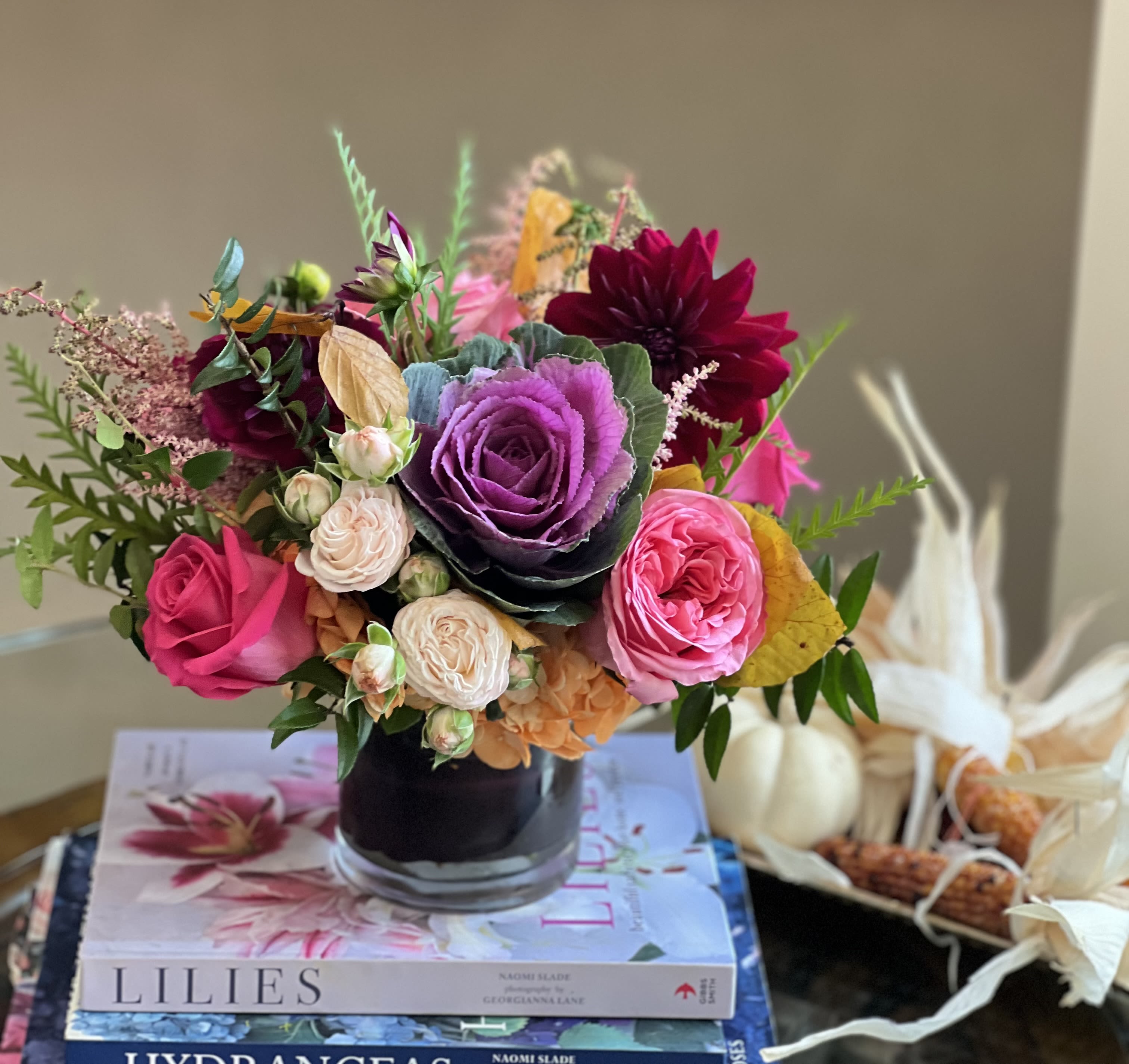 Maple Haze  Maple Haze  - Flowers: purple kale, pink expression garden roses, pink floyd roses, ivory bombastic spray roses, orange hydrangea, burgundy dahlias, and touches of grevillea.   Arranged in our 4x4 glass cylinder vase wrapped with a burgundy aspidistra leaf.