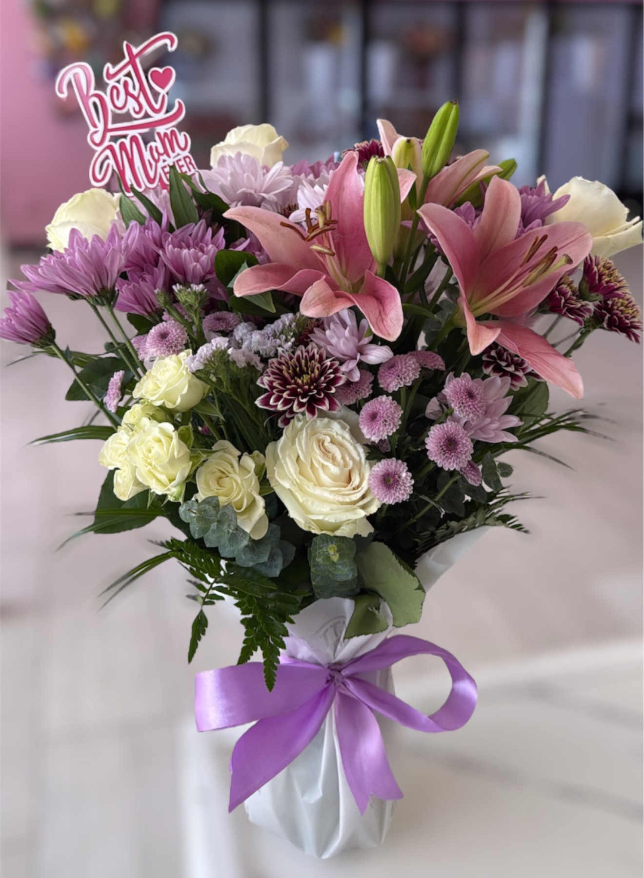 Sending Love Arrangement Sending Love Arrangement - A vase arrangement with a delightful mix of blooms. Light pink lilies stand tall among classic white roses and vibrant purple chrysanthemums, complemented by other assorted flowers that add texture and depth. A "Best Mom Ever" sign is nestled among the flowers, making it a perfect gift to show appreciation and love. Topper can be changed out based on occasion and note message (Anniversary, Birthday, Moms Birthday, Love You) Please note that florists may need to make substitutions based on inventory to ensure the freshest blooms are delivered, while maintaining the arrangement's overall aesthetic.