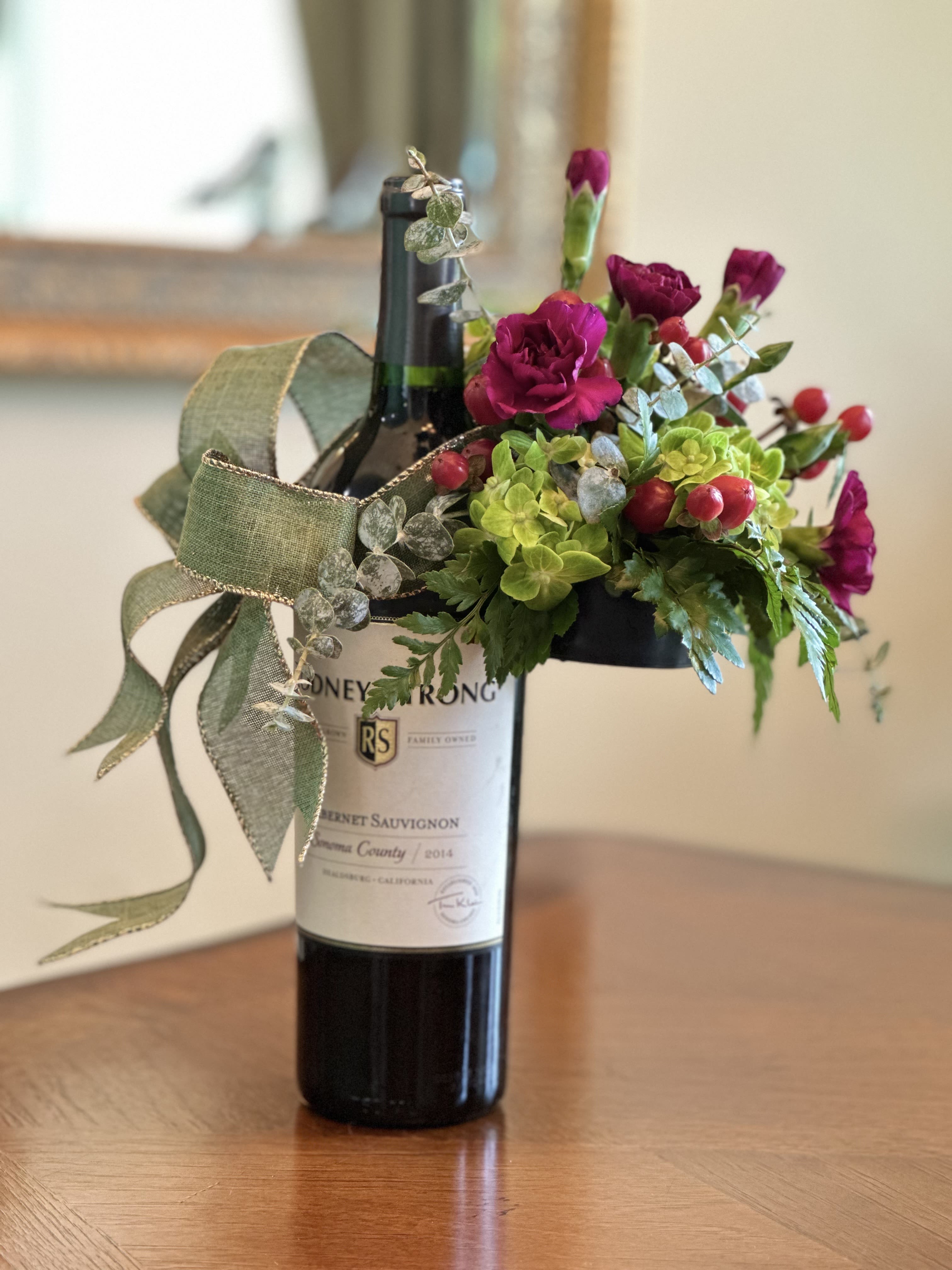 Vine &amp; Bloom Wine Clip - Add a touch of floral elegance to any bottle with our Vine &amp; Bloom Wine Clip. This handcrafted arrangement features rich magenta blooms paired with soft green hydrangea accents, lush foliage, and a hint of red berries. A graceful sage ribbon with gold trim completes the look, making it a perfect gift accent for wine lovers, dinner hosts, or special celebrations. Simply clip it onto your favorite bottle to transform it into a stunning floral presentation.  WINE BOTTLE NOT INCLUDED.