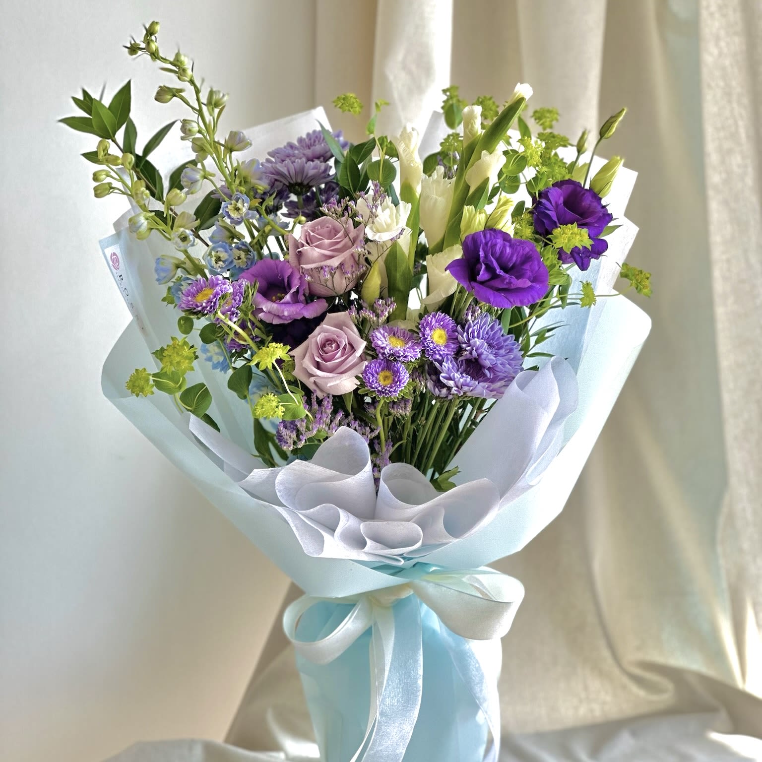 My Angel  - A front-facing bouquet with white, green, purple, and light blue flowers, wrapped in light blue paper.   Flower types may vary depending on the season.