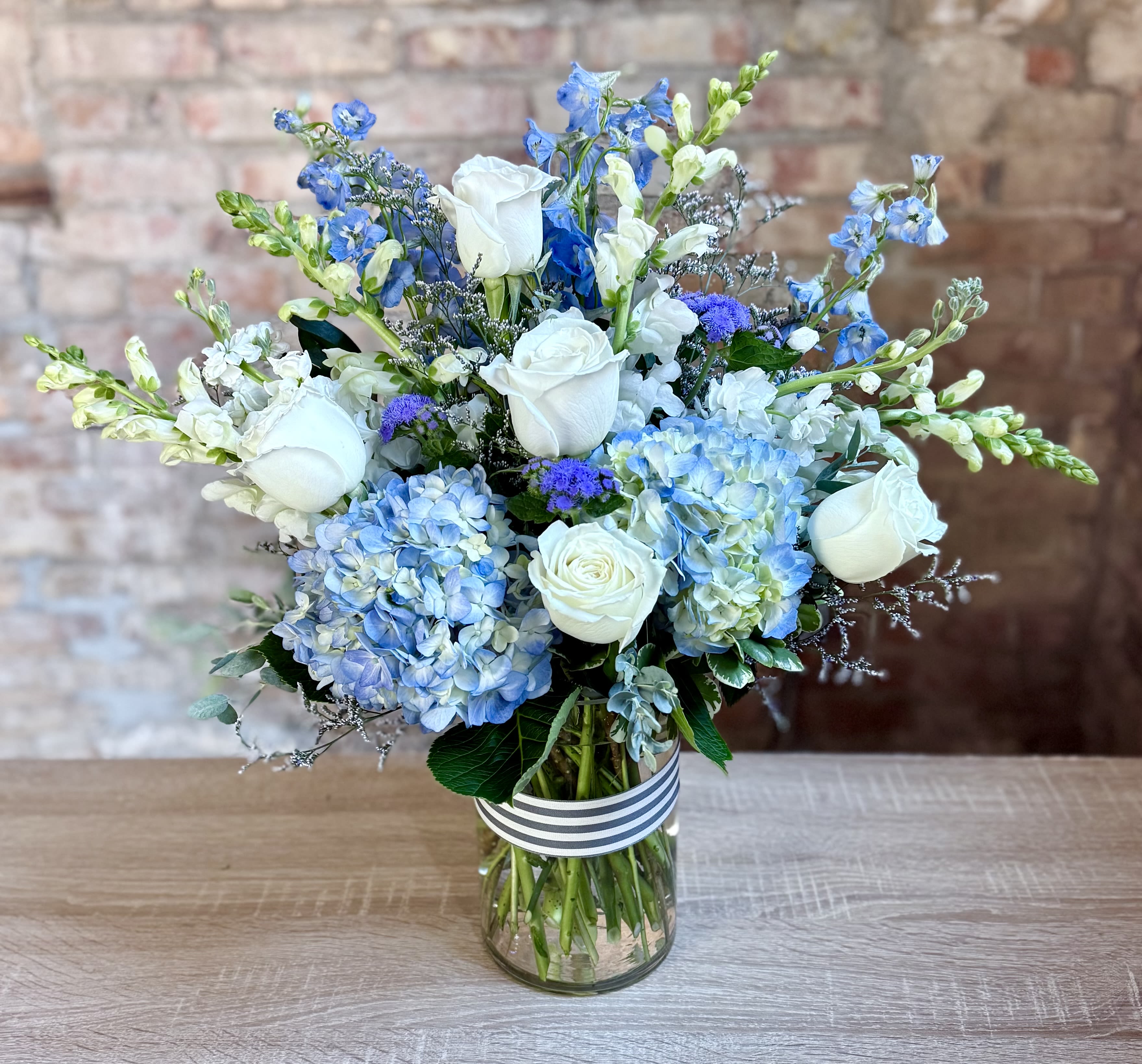 Pippa  - A clear glass cylinder filled with a collar of soft blue/periwinkle hydrangeas, white snapdragons, light blue delphinium, white roses, white stock, and lavender filler. Soft and pretty! 