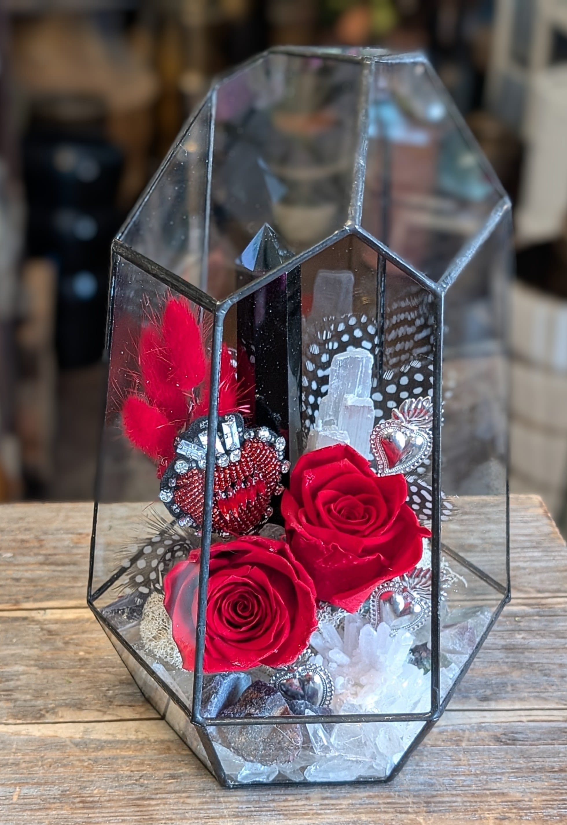 Bowie- Preserved Roses - Bowie by Windmill Floral Expressions in Las Vegas is a cosmic love relic—part altar, part art piece—designed for the bold, the mystical, and the beautifully untamed. Encased in a faceted glass terrarium, this arrangement transcends the ordinary, channeling glam-rock edge with sacred symbolism and romantic intensity.  At its core, two Preserved crimson roses burn with passion, their velvety petals glowing like embers in a crystal-strewn dreamscape. A powerful black obsidian crystal rises like a monument of strength, paired with a serene selenite shard that radiates light and clarity. Scattered silver heart charms catch the light like constellations, while a rhinestone-studded sacred heart, framed in fiery red bunny tails, pulses with divine energy—equal parts protection, passion, and punk devotion.  Delicate spotted feathers add softness and movement, whispering through the arrangement like fragments of stardust. Every element is intentional—raw yet refined, bold yet reverent.  Bowie is more than a floral arrangement—it’s a tribute to fearless love, iconic style, and soul-deep symbolism.  Available for same-day flower and gift delivery in Las Vegas and Henderson.