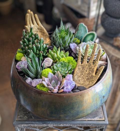 Mantra - Mantra is a meditative journey in botanical form—a lush sanctuary of grounding greens and sacred symbolism. This serene succulent garden features a curated mix of textures, from the architectural symmetry of zebra haworthia and jade to the soft, sculptural rosettes of echeveria and graptosedum. Sprinkled with vibrant moss, amethyst crystals, and polished stones, the piece hums with peaceful energy and natural beauty.  Golden hamsa hands rise like protective talismans among the foliage, their intricate patterns and spiritual meaning adding depth and intention to the design. Agate slices provide a rich contrast and a touch of elemental grounding, making Mantra not just a planter—but a space for presence, reflection, and beauty.  Perfect for home altars, mindful gifting, or simply bringing balance into any space. Delivered with care in Las Vegas and Henderson by Windmill Floral Expressions, where art and nature align in every creation.