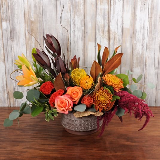 Modern Autumn Modern Autumn - This modern autumn bouquet of roses, orange lilies, proteas & magnolia leaves . Arranged in a keepsake container, it's a modern fall treat for any occasion!