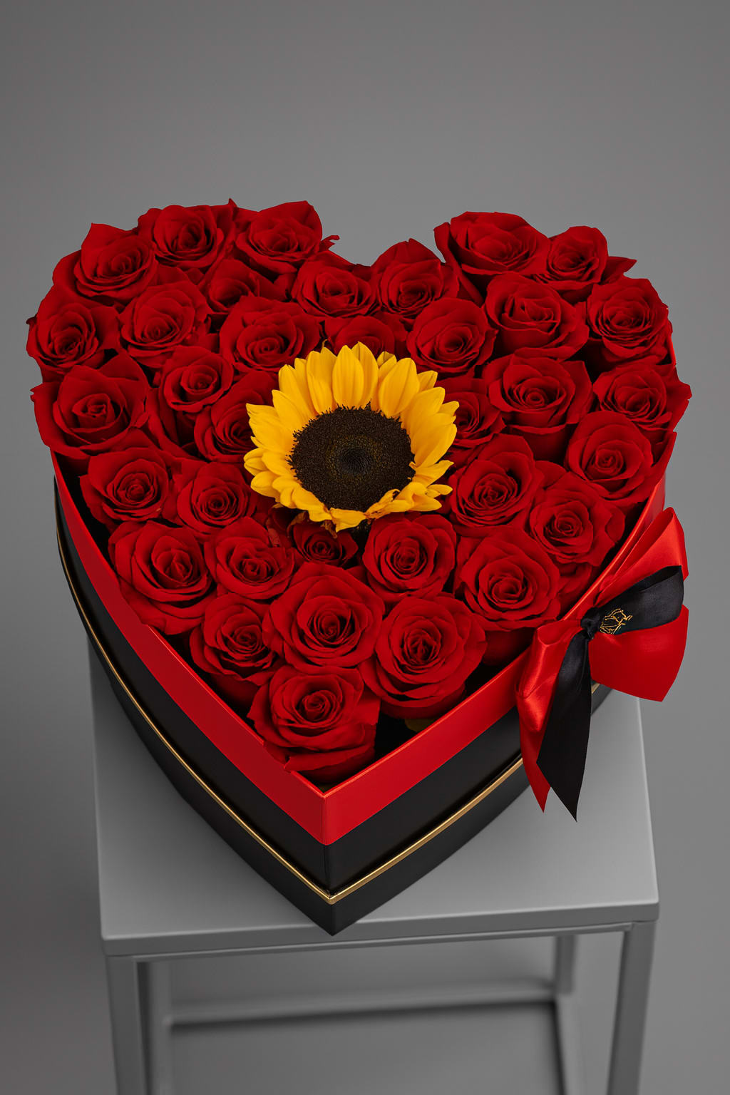 Sun-Kissed Box - Celebrate love with our Sun-Kissed Box, a stunning, heart-shaped arrangement of premium, hand-set roses symbolizing a heart overflowing with devotion. A single, vibrant sunflower bursts from the center, representing bright, enduring happiness.  Enclosed in a sleek black and red heart box and finished with an elegant ribbon, this arrangement is the ultimate statement gift for anniversaries, Valentine's Day, or any unforgettable moment.