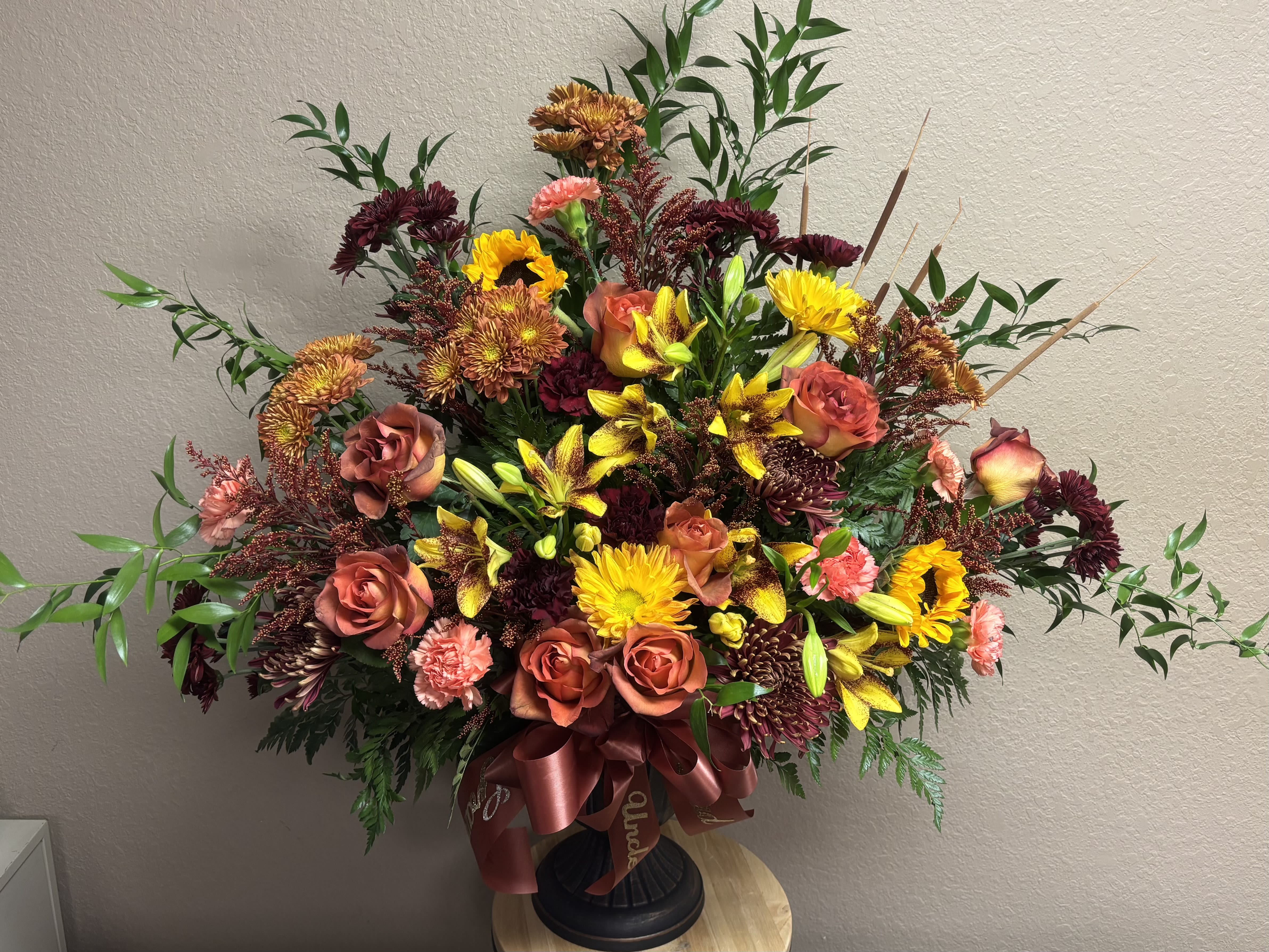 Autumn Radience Floral Arrangement  - Celebrate the warmth and beauty of the season with our Autumn Radiance Floral Arrangement. This stunning display features a harmonious blend of burnt orange roses, golden yellow lilies, rust-colored chrysanthemums, burgundy mums, and soft coral carnations, all nestled among lush greenery and accent foliage. Designed in a classic pedestal vase and finished with an elegant satin ribbon, this arrangement captures the rich tones and textures of fall — perfect for a heartfelt sympathy tribute.