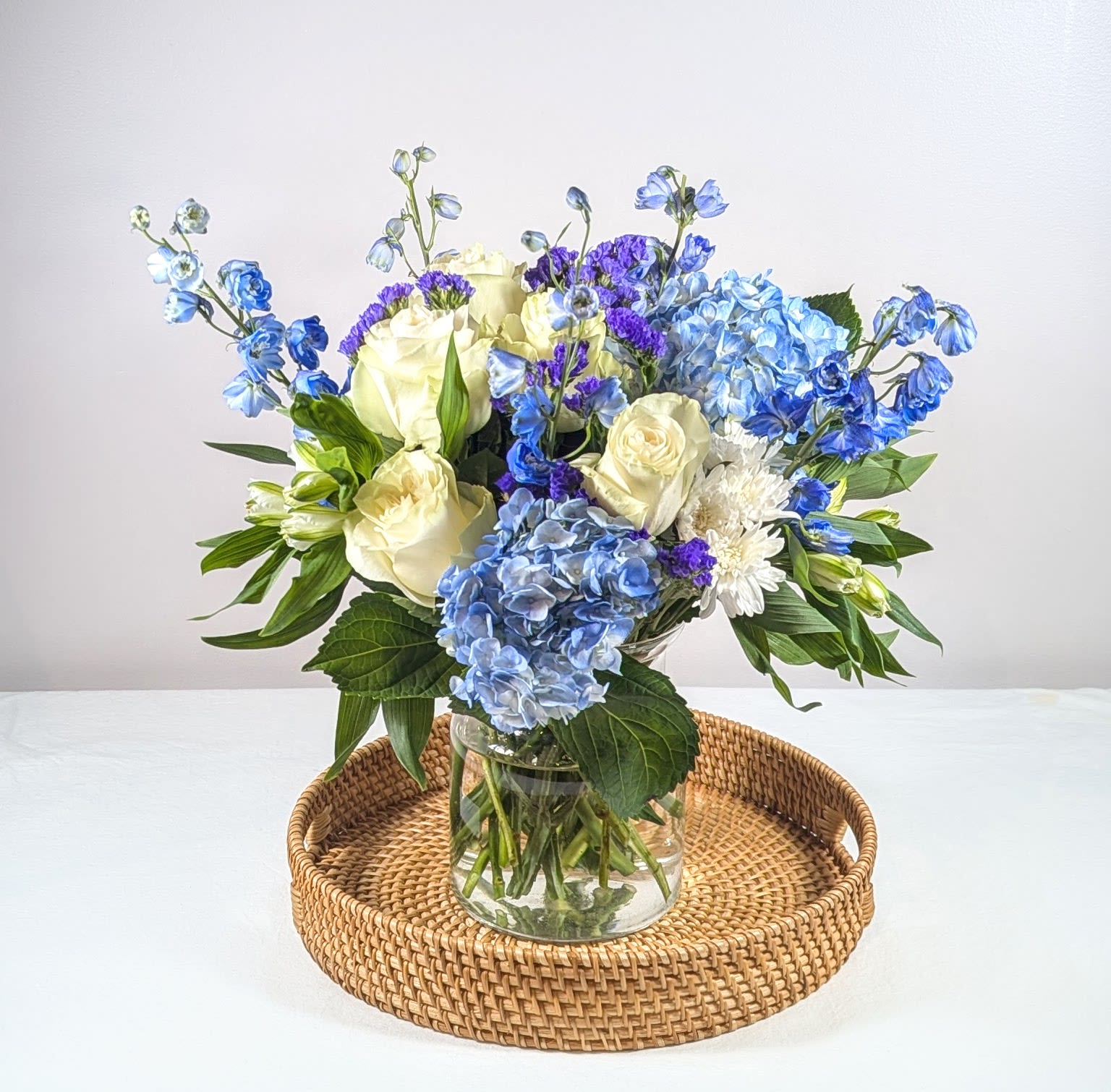 Blue Heaven - A gorgeous and dazzling blue and white arrangement with delicate blue belladonna delphinium, white roses, bunches of purple statice, blue hydrangeas, white poms, white alstroemeria &amp; purple thistle, accented with dark greenery. Arranged &amp; delivered in a clear glass vase.    Measures approx. 20&quot; tall and 18&quot; wide Orientation: All Around