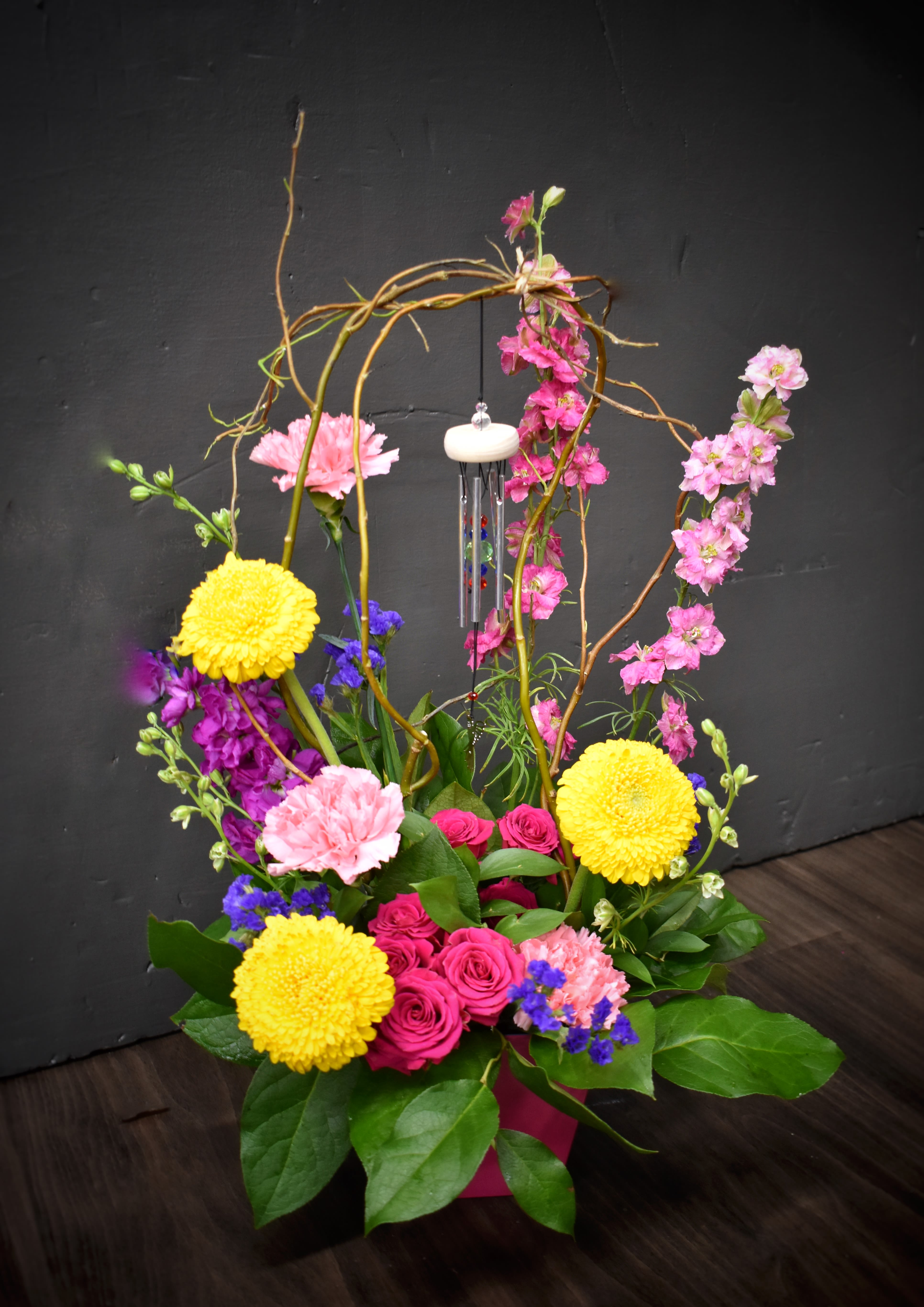 Melody Blooms Melody Blooms - An arrangement to enjoy with your eyes, nose AND ears, this piece features a colorful mix of larkspur, fragrant stock, bright daisies, spray roses and carnations settled in a matching reusable container.  This arrangement speaks volumes with delicate mini chimes hanging beneath an arch of willow branches.