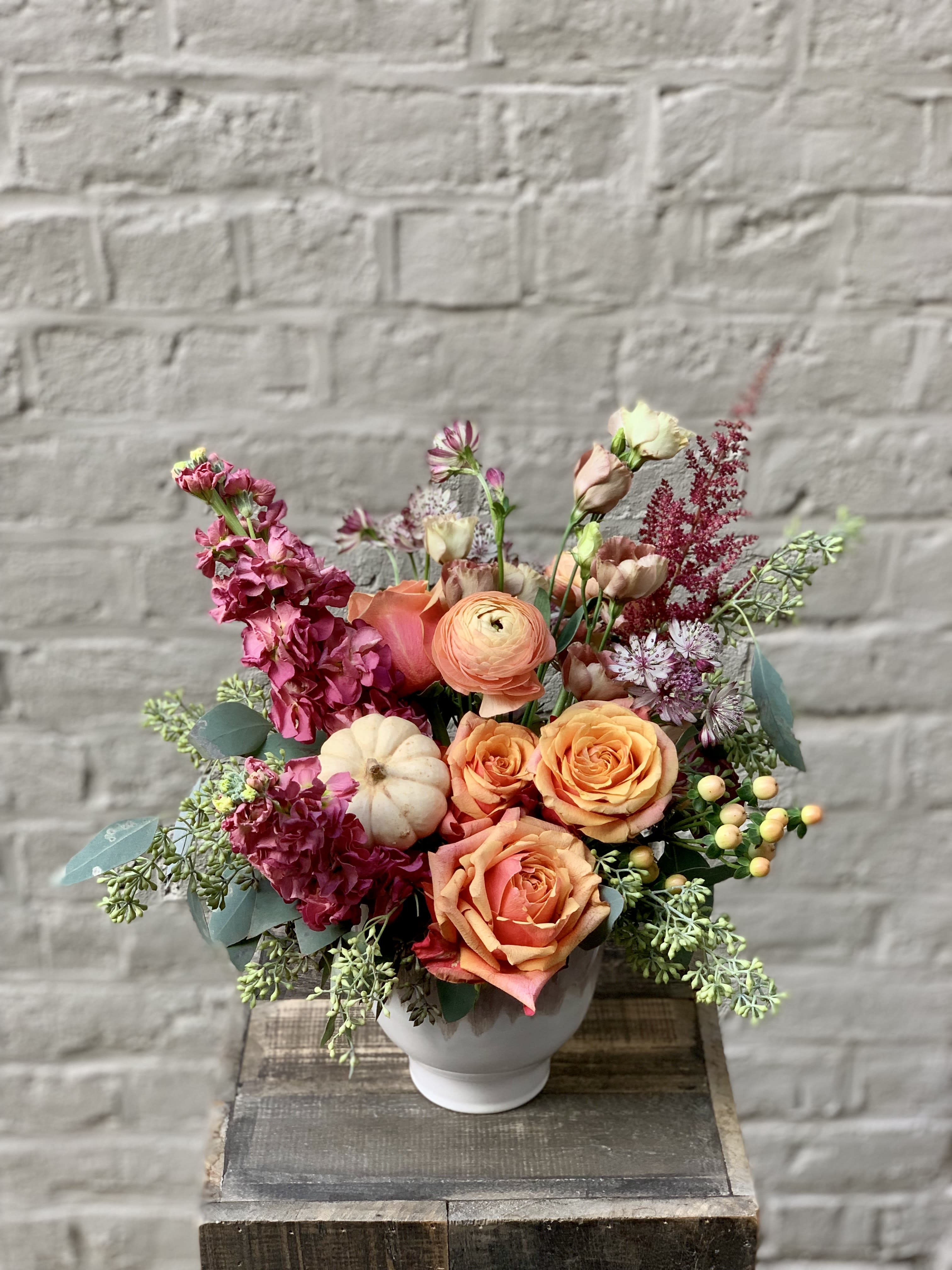 Autumn Embrace Autumn Embrace - Inspired by the inviting warmth of a tender embrace, this elegant arrangement is filled with delights that will keep you cozy through the chill that the autumn air brings. Roses, ranunculus, hypericum berries, the whimsical touch of locally-grown white pumpkin, and more fill a unique two-tone ceramic pedestal vase. Arrangement measures approximately 11" wide by 14" tall.