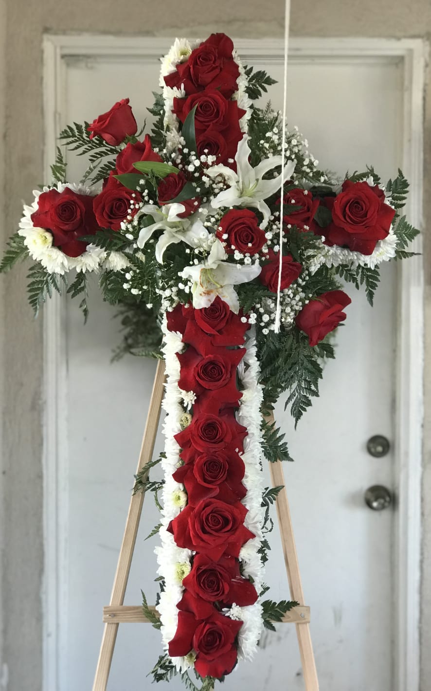 Eternal Love Cross- Red - This elegant funeral cross features a striking row of red roses cascading down the center, symbolizing deep love and remembrance. A serene white outline of lilies and cushion poms adds purity and grace, creating a heartfelt tribute to honor the memory of a loved one.    Note: PICTURED IS THE DELUXE SIZE.  Easel is INCLUDED