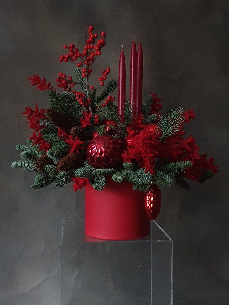 Red Velvet Christmas - Capture the warmth and wonder of the holiday season with Crimson Noel Glow, a breathtaking arrangement styled in rich tones of deep red and lush evergreens. Elegant taper candles rise from the center, creating a radiant focal point surrounded by fluffy red blooms, festive ornaments, natural pinecones, and seasonal greens. Designed in a matte red cylinder container, this arrangement brings drama, luxury, and classic Christmas charm to any table or mantle.