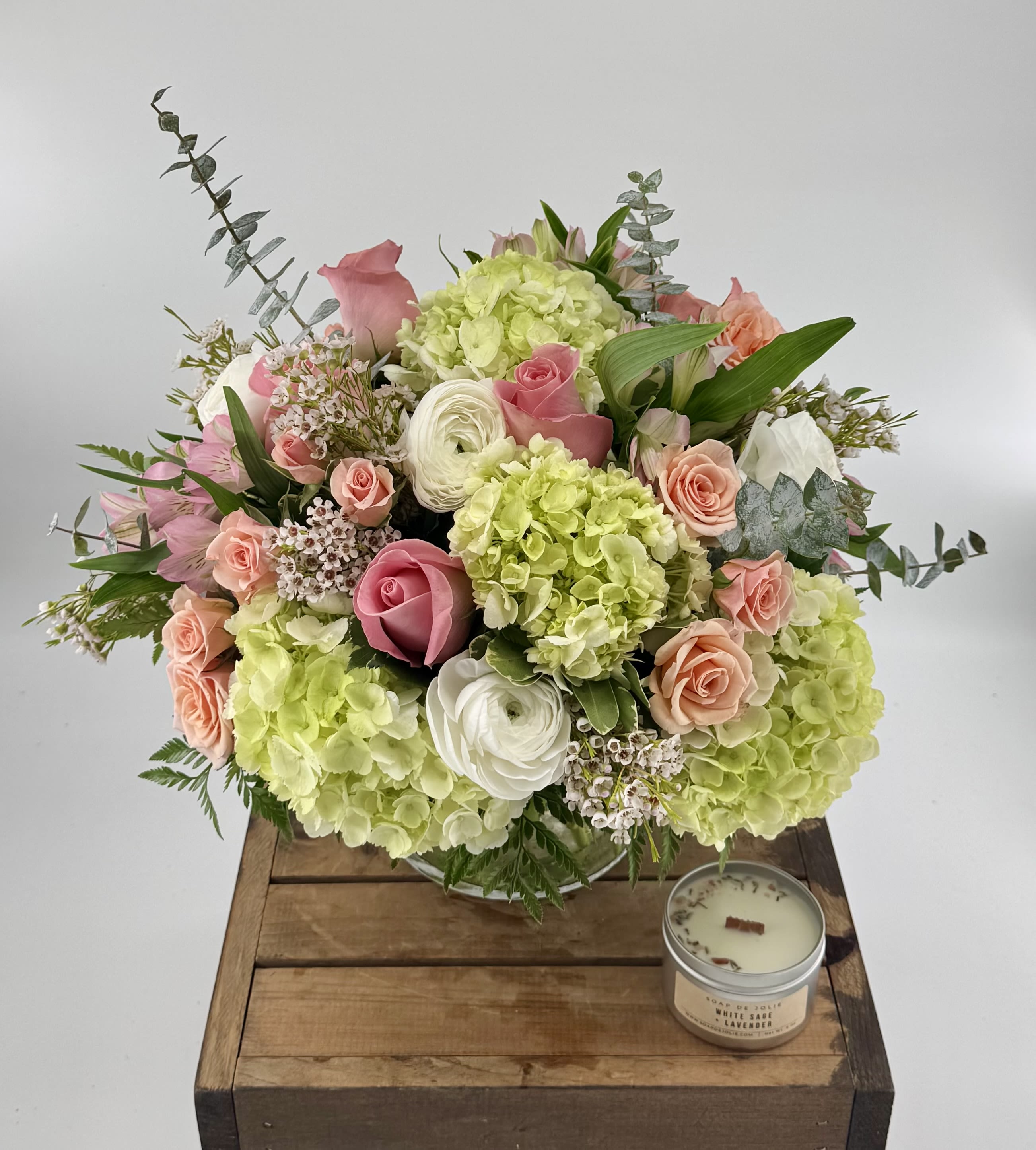 Simplicity  (BB) - Delight in a stunning blend of green and white hydrangea, white ranunculus, pink roses, white alstroemeria, pink spray roses, wax flower and eucalyptus, artfully arranged in a leaf-lined 6x6” clear vase—crafted to elevate your most cherished moments. 