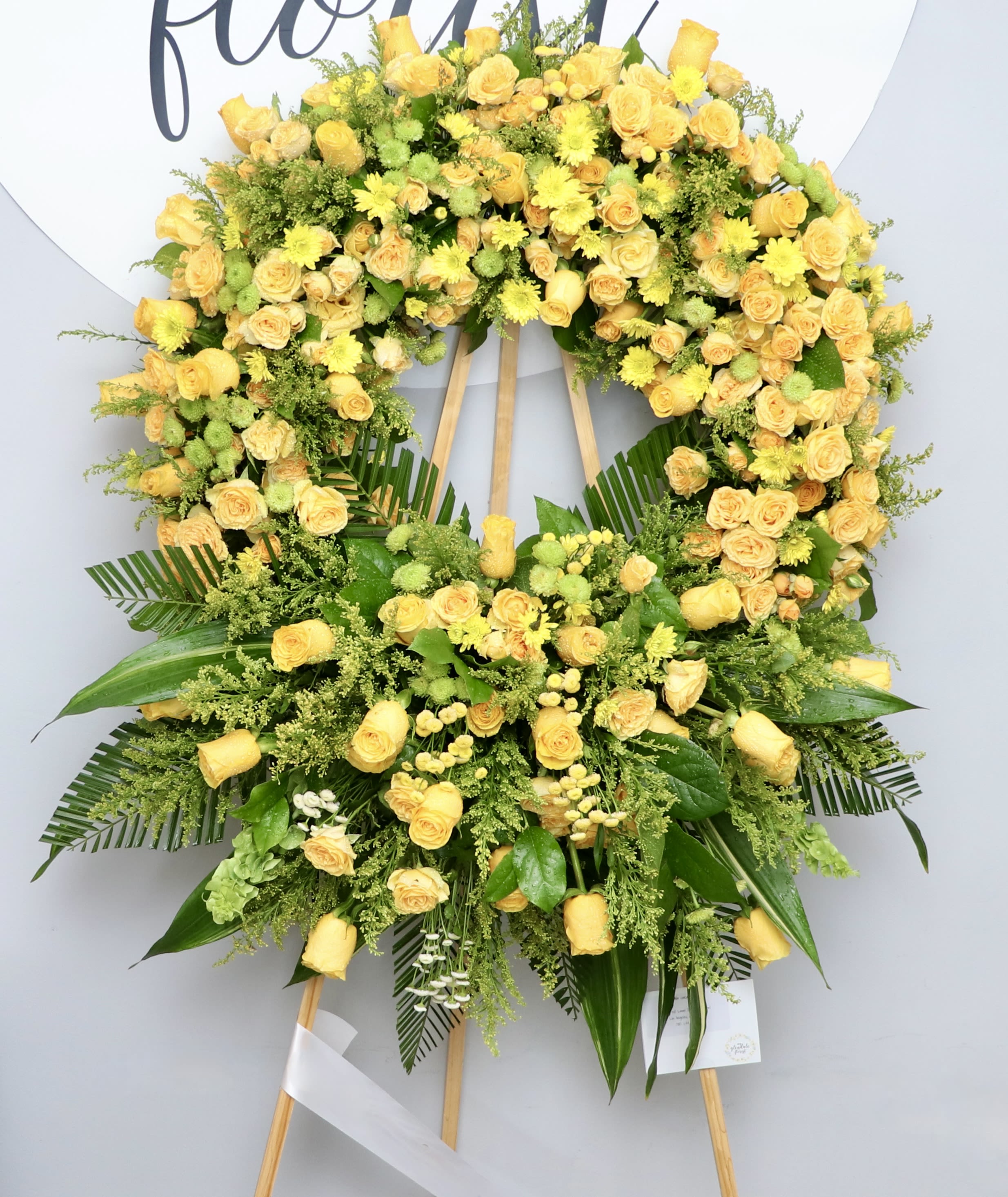 Golden Horizon  - The Golden Horizon is an elegant funeral wreath, featuring yellow roses and seasonal greenery. The combination of colors and textures create an inviting and comforting atmosphere that celebrates the life of the departed.  We include easel, printed banner, and delivery (some fees may apply). An additional banner can be added for $15  Standard size is 30'', deluxe is 36'', and premium is 42''. 
