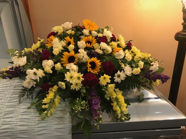 Golden Memories Casket Spray - A radiant tribute of warmth and remembrance, the Golden Memories Casket Spray features cheerful sunflowers surrounded by soft white roses, yellow snapdragons, creamy carnations, and deep burgundy blooms. This uplifting display celebrates a life well lived, blending beauty and comfort in every petal. Perfect for honoring a loved one with the brightness and joy they brought to those around them.