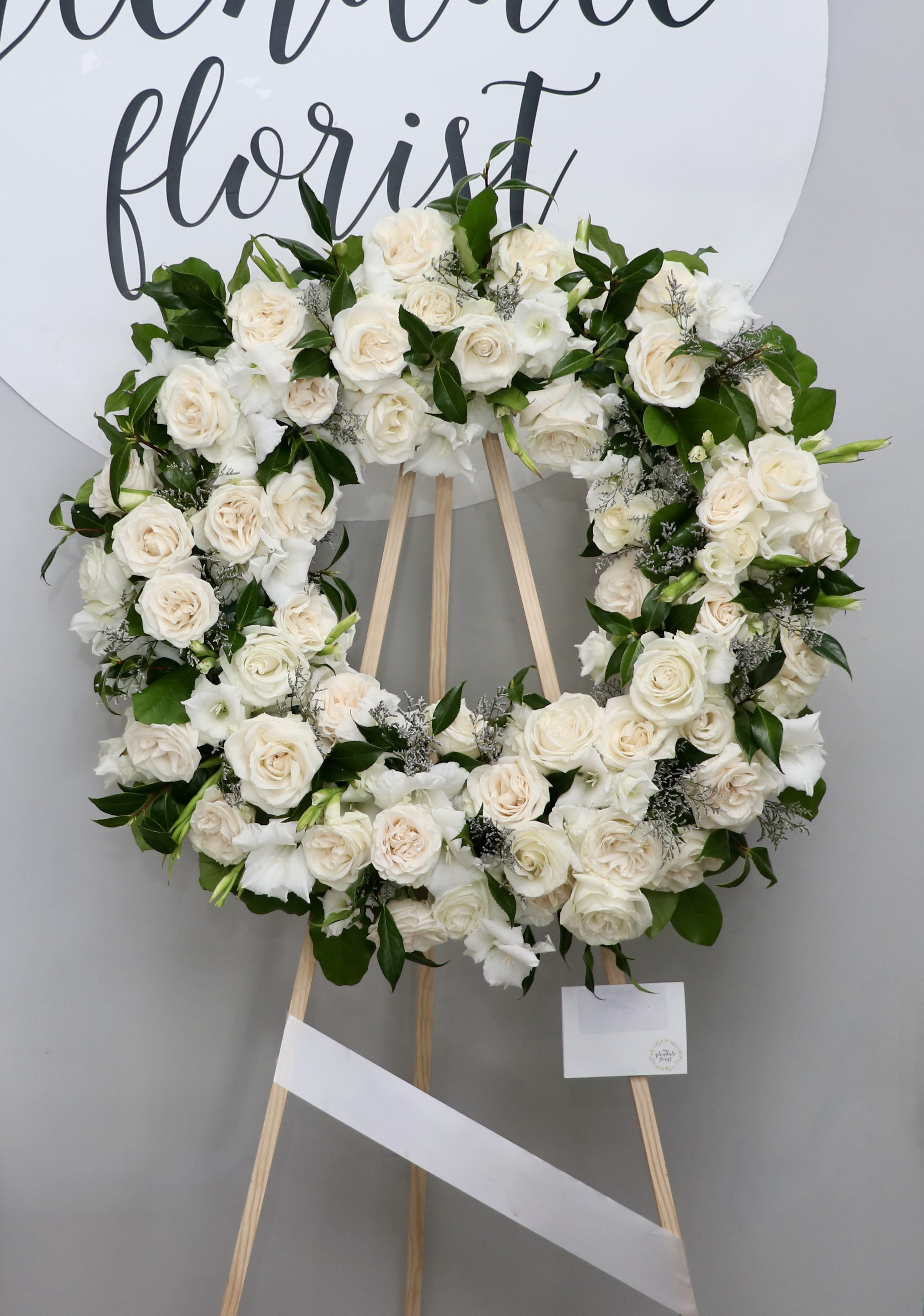 Ivory Grace Wreath  - This sympathy wreath is elegantly designed with white roses, gladiolus, and greenery.  We include easel, printed banner and delivery (some fees may apply). An additional banner can be added for $15  Standard size is 30'', deluxe is 36'', and premium is 42''.