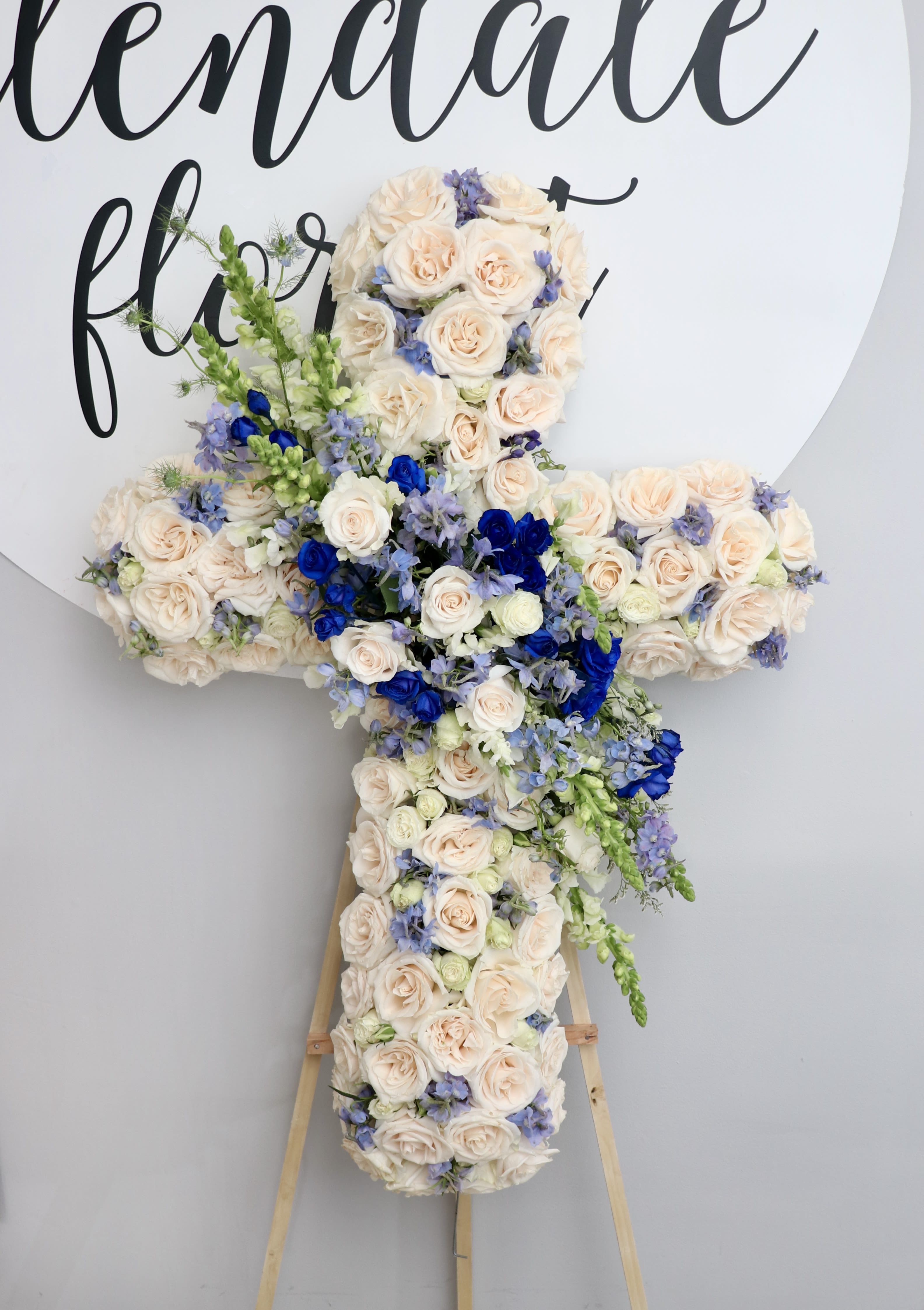Blue Dream Cross - This sympathy cross is made with white roses and accented with shades of blue. This cross is a lovely choice if you are looking to add some color to your loved one's service.   We include easel, printed banner and delivery (some fees may apply). An additional banner can be added for $15.  Standard size is 30'', deluxe is 36'', and premium is 42''.