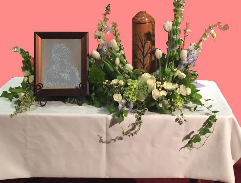 A Touch Of Color Remembrance Arrangement - Our exquisite “S-Shaped” remembrance arrangement is inspired by the lasting impressions that a loved one makes on our lives. Remembrance arrangements are hand-crafted to surround an urn or curve gently around a photograph and urn together, as in these photos.  Every one of these arrangements are as unique as the loved one it honors.  While they are available with any color accent**, it will always feature premium flowers professionally designed in various heights to accent an urn, photo, or candle.  This is a one-of-a-kind arrangement. While the tulips in this arrangement are seasonal, most of the other flowers are available year-round, such as: roses, spray roses, hydrangea, Queen Anne’s lace, bells of Ireland, and larkspur. These lovely blooms are nestled in with a variety of premium greenery.  **Please specify your accent color in the special instructions. If you do not choose a color, this design will be made in all white.  