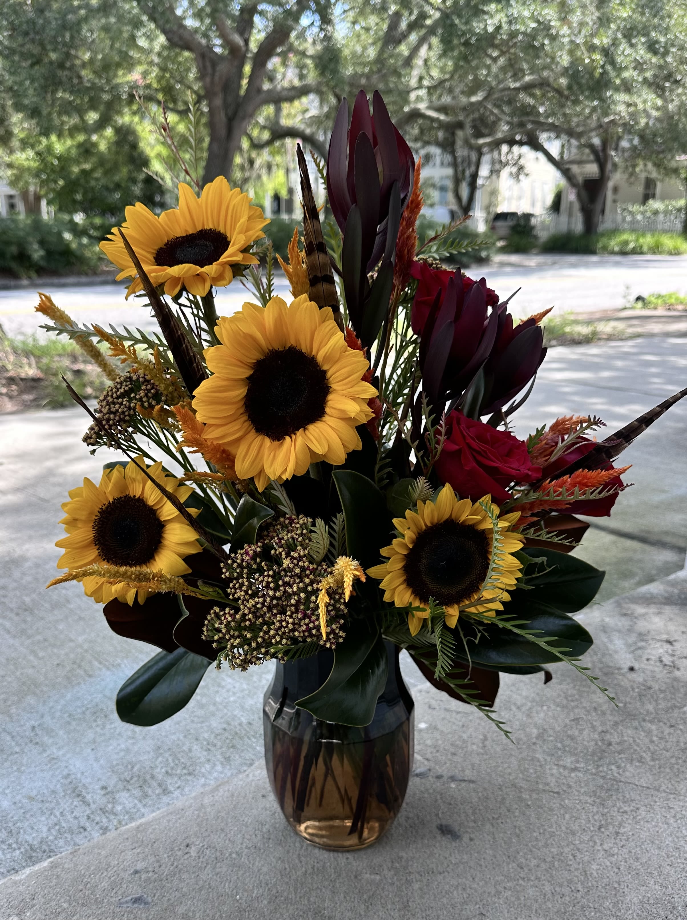 Feather & Flame Feather & Flame - Ignite the season with our Feather & Flame arrangement, inspired by the fiery spirit of The Hunger Games. Bold golden sunflowers and crimson roses burn bright against dramatic burgundy foliage, accented with pheasant feathers that nod to Katniss's woodland roots. A striking mix of warmth and strength, perfect for anyone ready to embrace their inner "Girl on Fire". *Photo shows a deluxe size*