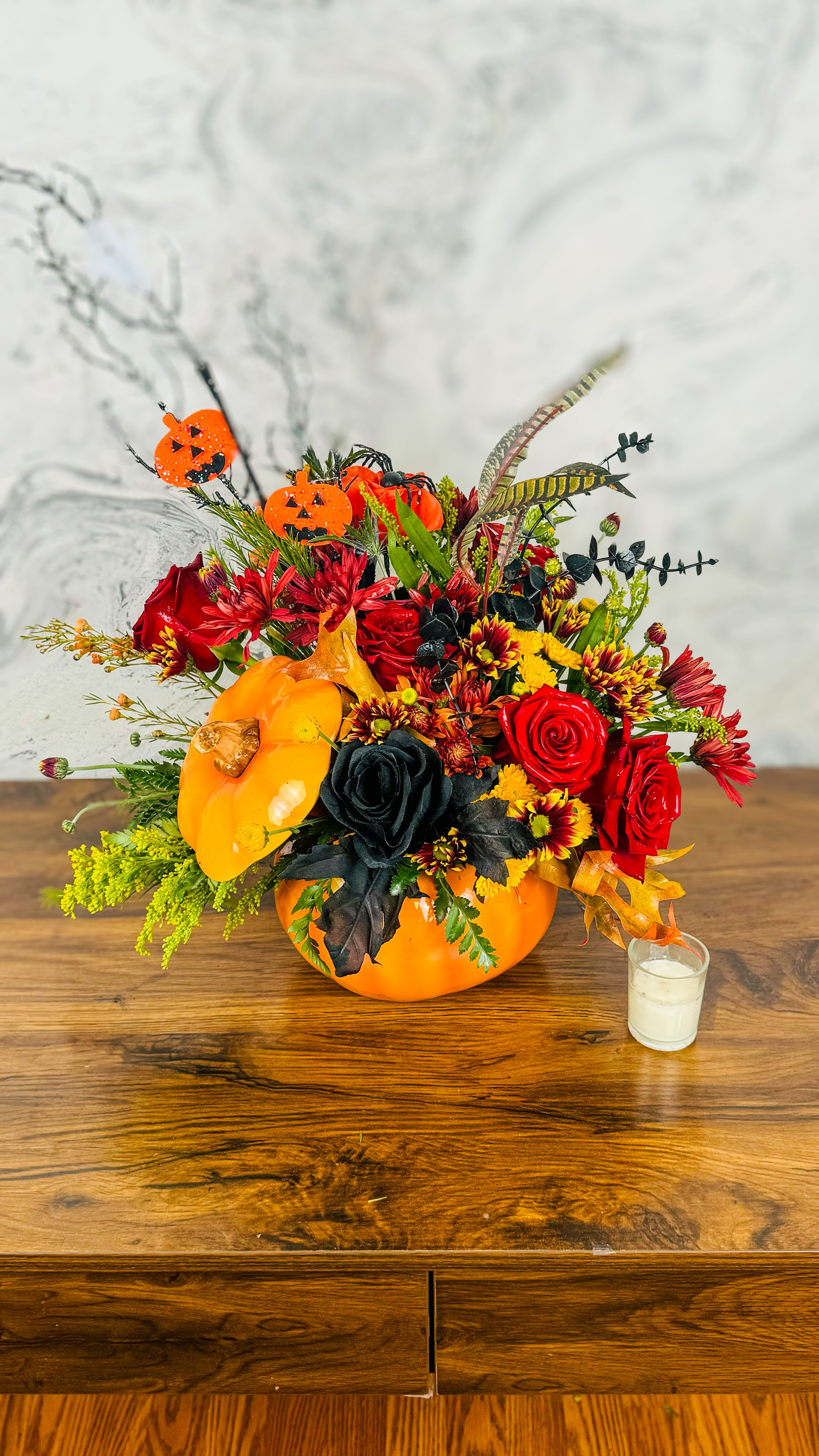 Halloween  Harvest - Halloween Harvest&quot; is a vibrant arrangement that captures the essence of autumn's beauty. Nestled within charming pumpkin containers, a delightful mix of fall-colored flowers—rich oranges, deep reds, and sunny yellows—brings warmth and cheer. Adding a whimsical touch, artificial Hallowin roses intermingle with the blooms, creating a festive tribute to the season. Perfect for enhancing your fall décor, this arrangement embodies the spirit of harvest time and the joy of Halloween.