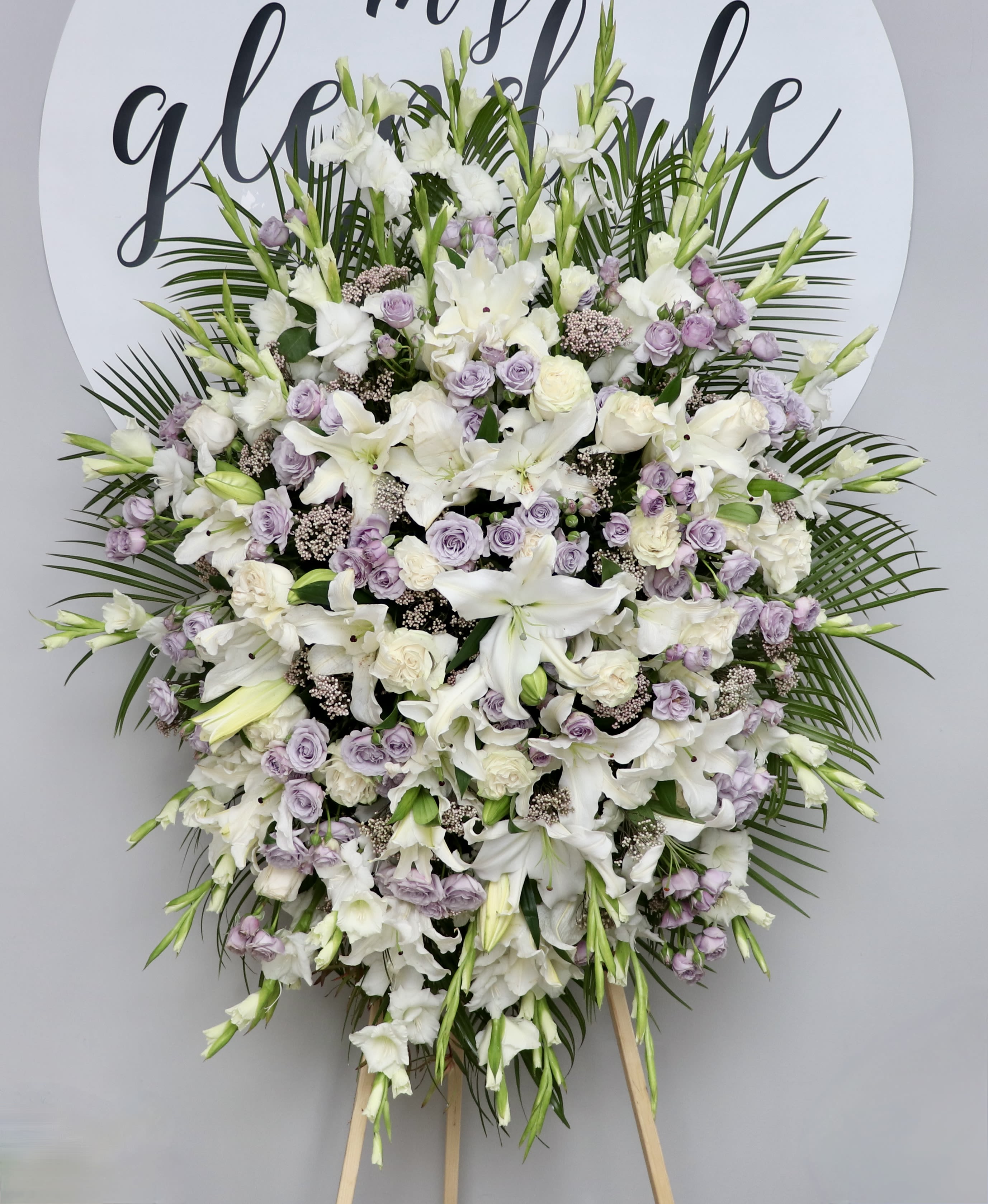 Wings of Lavender  - This spray contains a collection of bright lavender roses and white lilies surrounded by seasonal greens and gladiolas.  We include easel, printed banner, and delivery (some fees may apply). An additional banner can be added for $15.  Standard size is 30'' wide, deluxe is 36'', and premium is 42''.