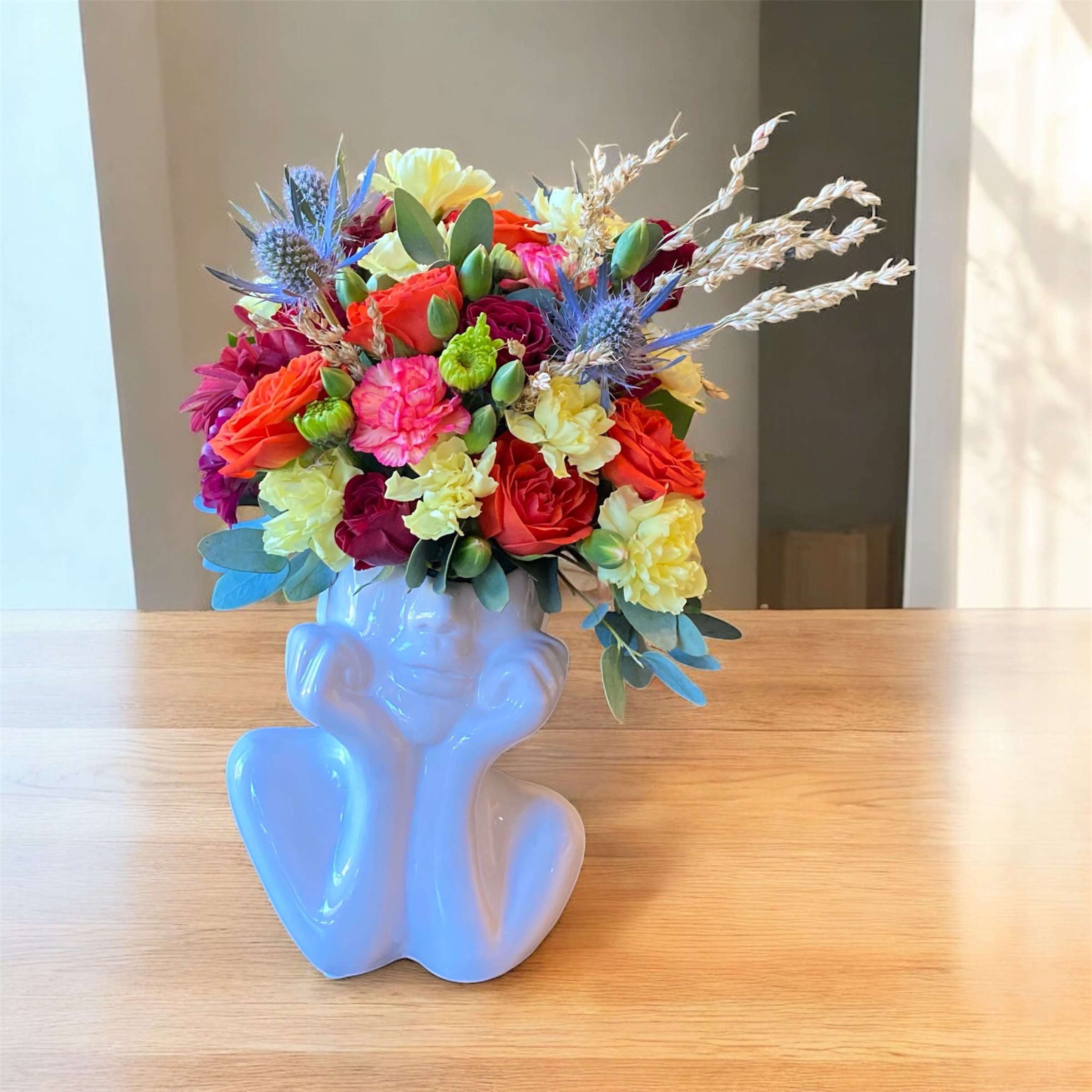 Bloom thought  Bloom thought  - A white sculpted face adorned with autumn blooms. Red, orange, and yellow flowers bring warmth and life to this elegant arrangement. A celebration of fall’s vibrant beauty.