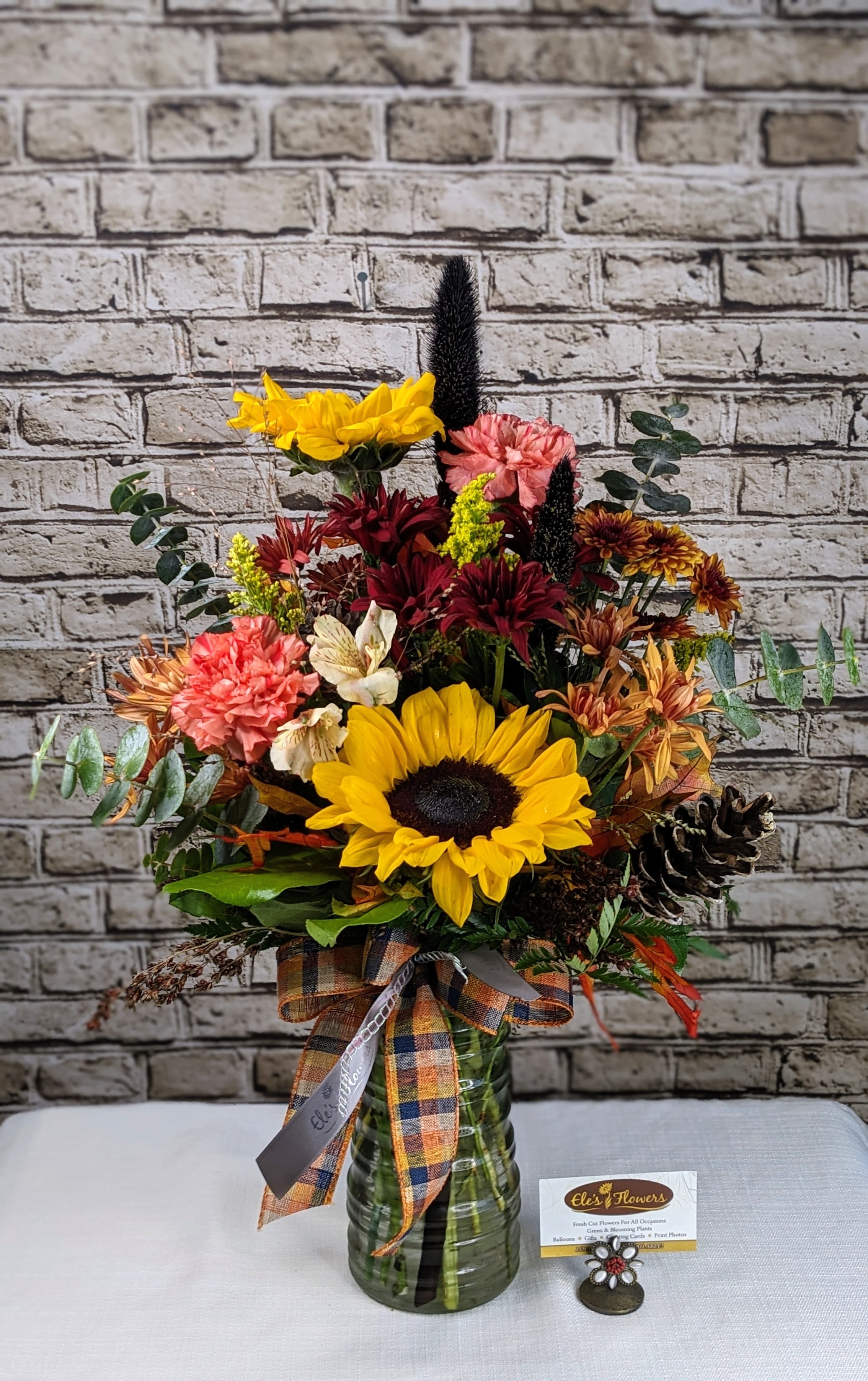 Autumn Fields Autumn Fields - Celebrate the beauty of the season with this tall, textured arrangement bursting with warm autumn tones and rustic charm. Designed in a ribbed green glass vase and finished with a plaid ribbon bow, this piece brings a cozy, countryside feel to any space. Perfect for a fall centerpiece, a thoughtful gift, or a cheerful accent for your home.   The designs we create are custom and the blooms and containers will vary. We use the freshest product available to create the perfect floral combination for the recipient. If you have any special requests, please add those in the "special instructions" portion of your order to help us dial in your vision. Otherwise, our skilled designers, Sara & Brett, will take it from here!  *Please note that the photos are examples only – floral selection will vary.*  Approximate dimensions: 24" tall x 16" wide.