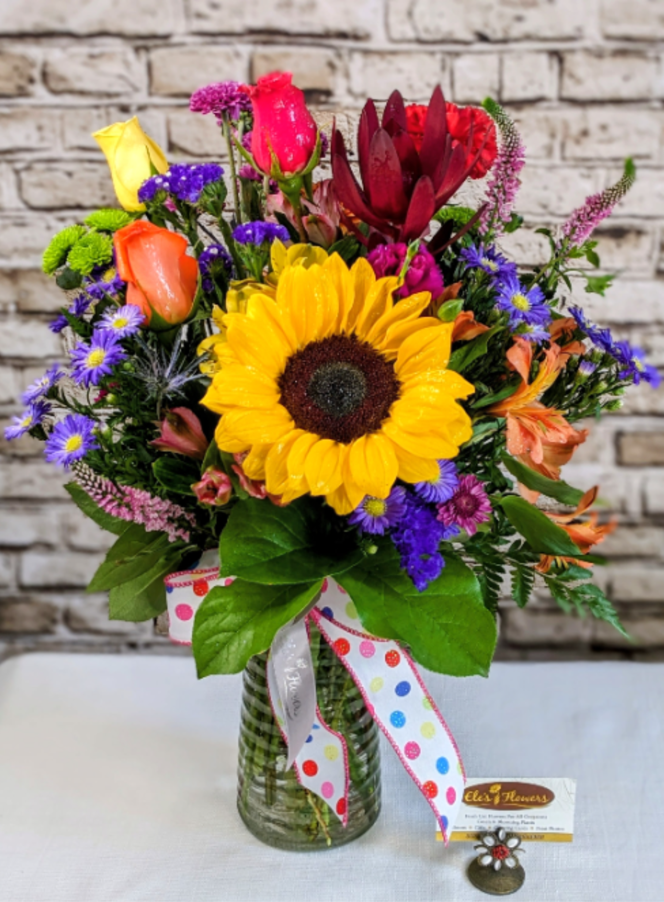 Bright Fall Cheer Bright Fall Cheer - Bring sunshine and smiles to their special day with this vibrant, joy-filled design. Bursting with bold color and playful texture, this arrangement captures the happy energy of fall celebrations. Designed in a clear glass vase and finished with a festive polka-dot ribbon, it’s the perfect way to brighten a birthday or simply make someone’s day unforgettable.  The designs we create are custom and the blooms and containers will vary. We use the freshest product available to create the perfect floral combination for the recipient. If you have any special requests, please add those in the "special instructions" portion of your order to help us dial in your vision. Otherwise, our skilled designers, Sara & Brett, will take it from here!  *Please note that the photos are examples only – floral selection will vary.*  Approximate dimensions: 22" tall x 15" wide.