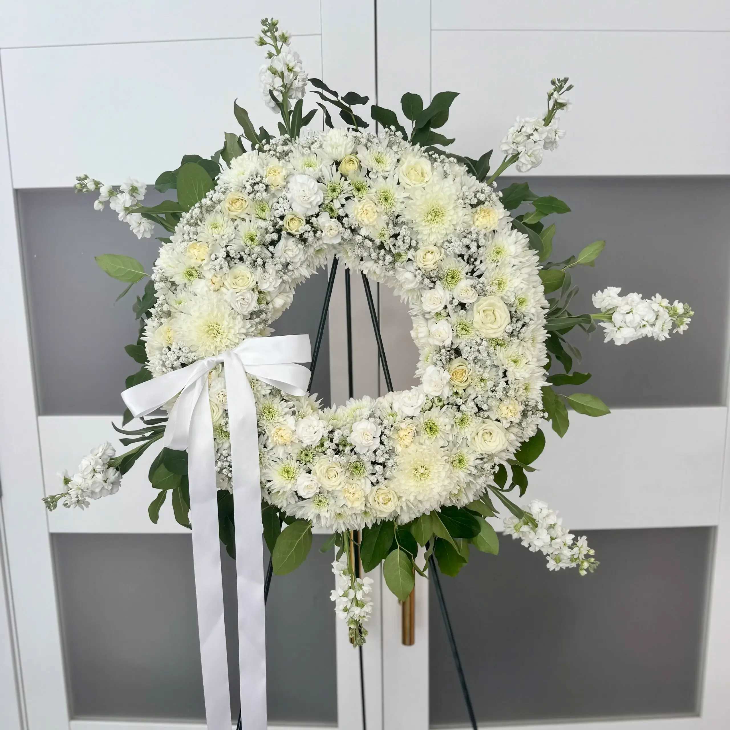 Funeral Wreath - Show your condolences with this elegant all white wreath which includes premium flowers and greenery accent  Colors could be changed as needed  * Easel Stand is included  Sizes: Medium (Standard) Large (Deluxe)