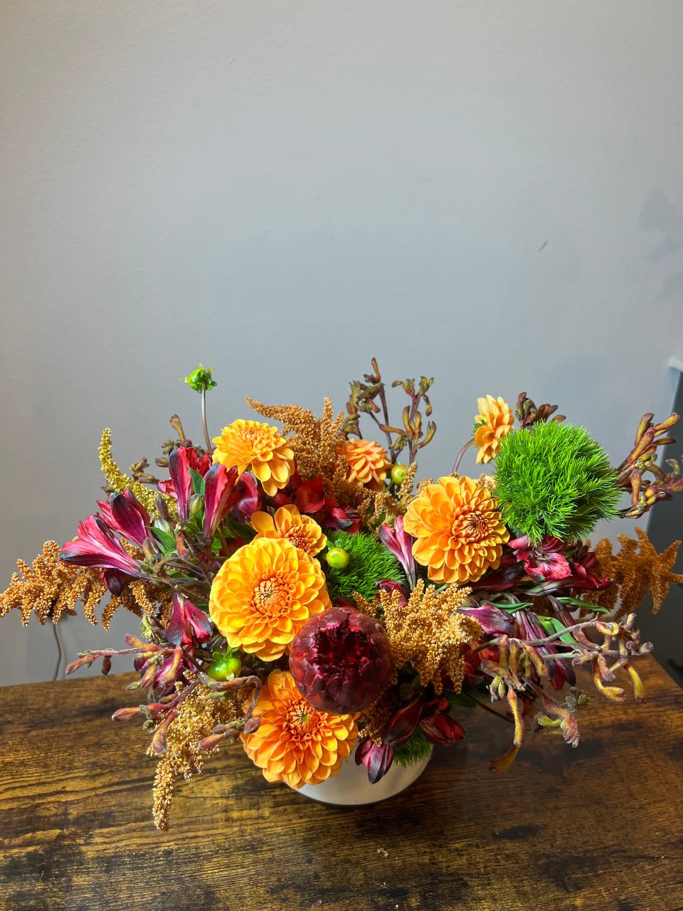 Autumn Glow — A Burst of Warm Elegance - Autumn Glow — A Burst of Warm Elegance  A lush arrangement inspired by the golden hues of autumn. Featuring vibrant orange dahlias, rich burgundy accents, and touches of green moss for contrast, this piece captures the cozy warmth and beauty of the season.  Perfect as a centerpiece for gatherings, a thoughtful gift, or a way to bring fall’s magic indoors. Each stem is artfully arranged to create a natural, yet refined balance of texture and color.  Details:  Fresh seasonal blooms in orange, burgundy, and green tones  Arranged in a modern ceramic vase  Handcrafted with care by Bluebell Flowers