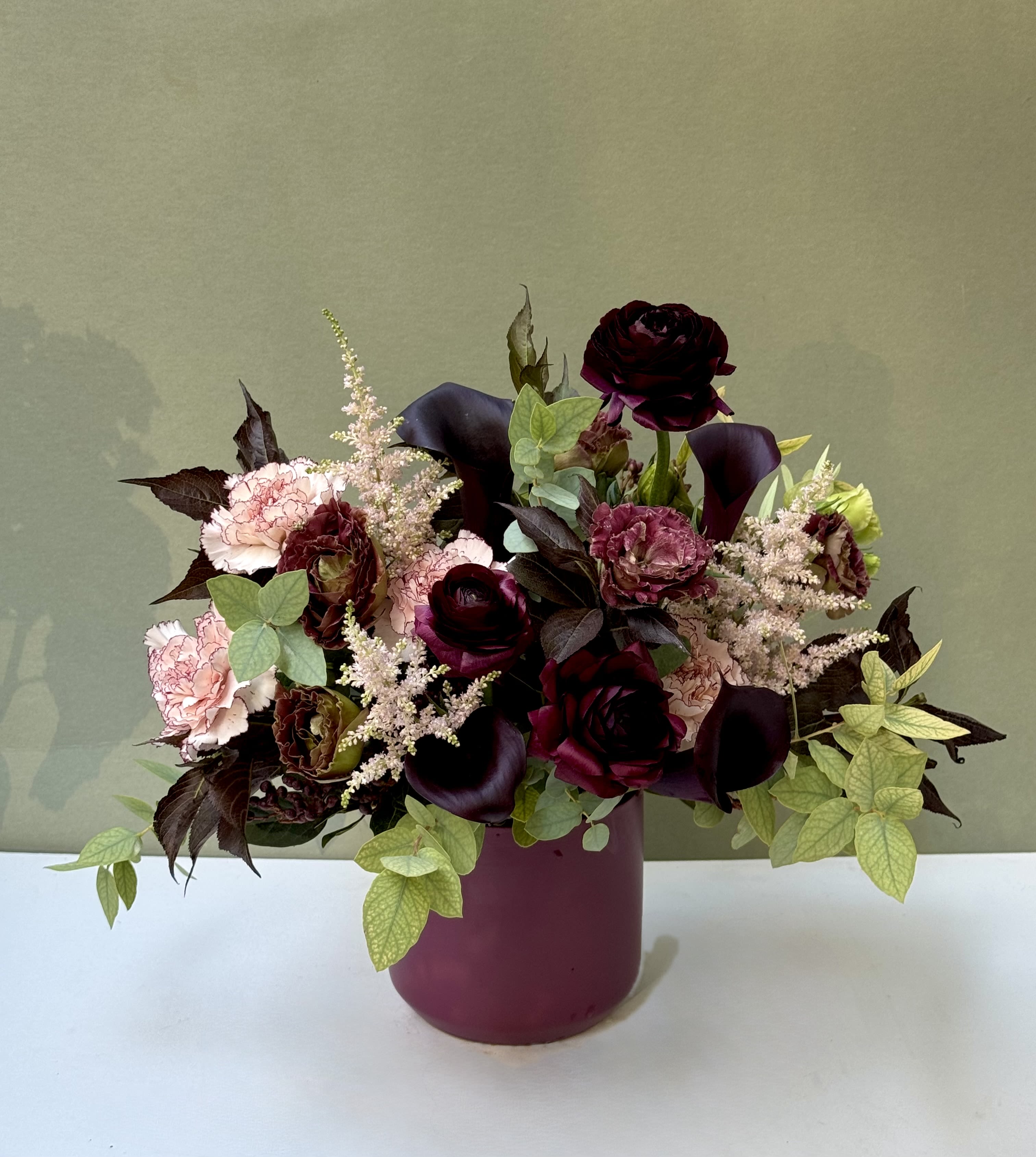 Velvet Noir  - Sophisticated and sultry, Velvet Noir is a stunning blend of deep burgundy ranunculus, calla lilies, and ruffled carnations paired with soft blush astilbe and fresh green foliage. Arranged in a matte plum vase, this design exudes timeless elegance with a modern edge. Perfect for romantic gestures, dinner parties, or adding a dramatic touch to any room, Velvet Noir is the definition of understated luxury.