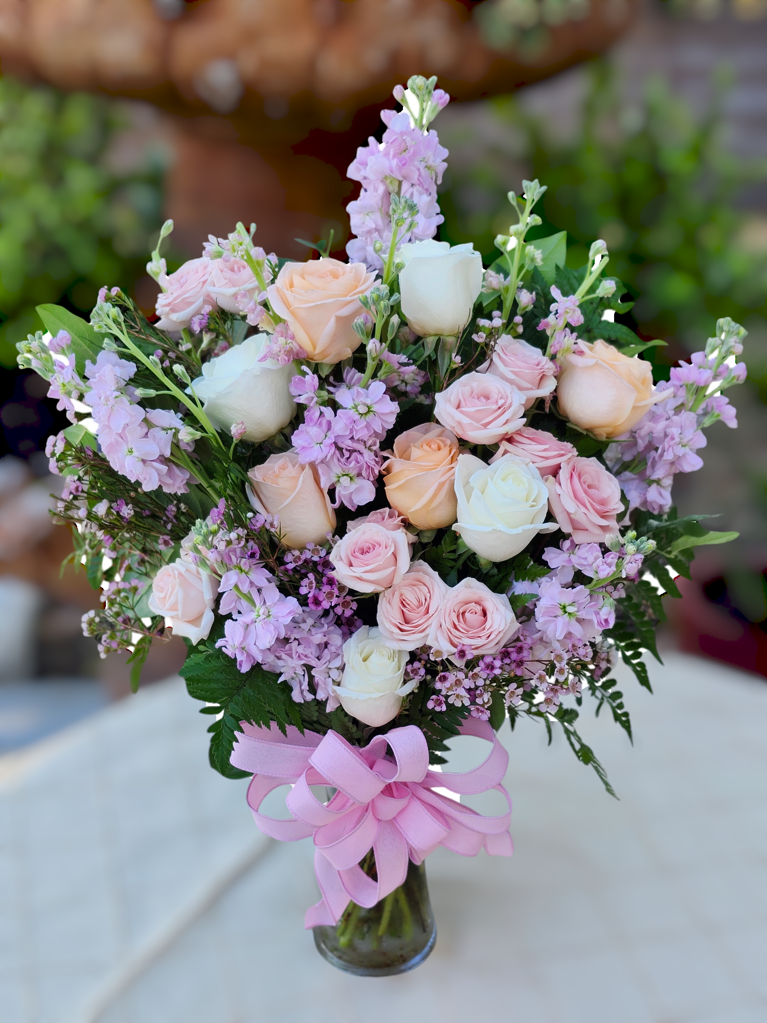 Soft Blush - Tall soft colors lavender, peach, pinks one sided vase. With a beautiful full bow. Stock, Roses, Wax, colors and flowers may vary due to inventory and season. 