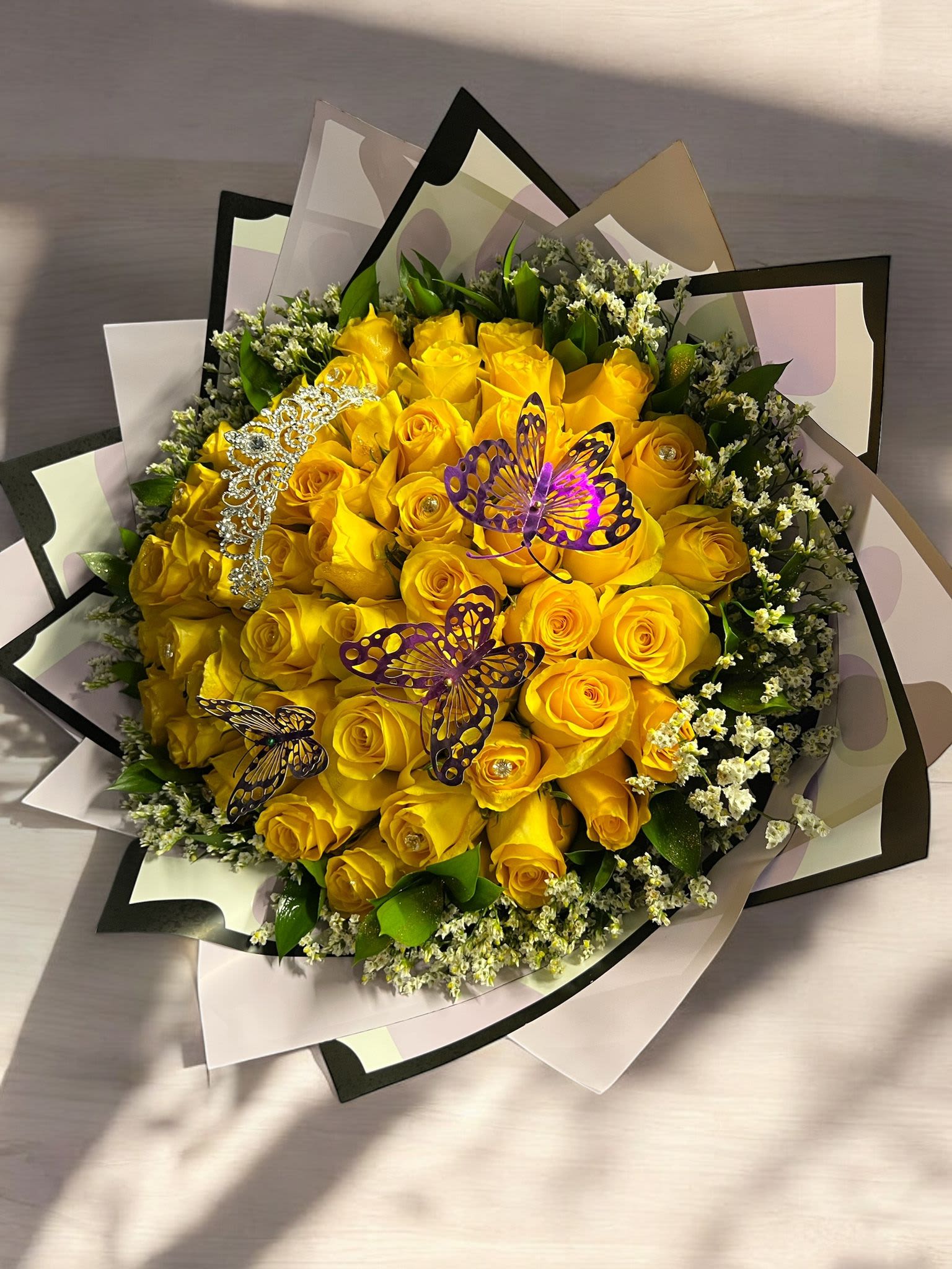 BuchunJul28 - Radiant Yellow Rose Bouquet Brighten any occasion with this stunning bouquet of fresh yellow roses, beautifully arranged and accented with delicate white filler flowers and lush greenery. Adorned with sparkling embellishments and elegant butterfly decorations, this bouquet is a perfect symbol of joy, friendship, and celebration. Wrapped in stylish paper with a modern design, it makes a thoughtful gift for birthdays, anniversaries, congratulations, or simply to bring sunshine to someone’s day.    