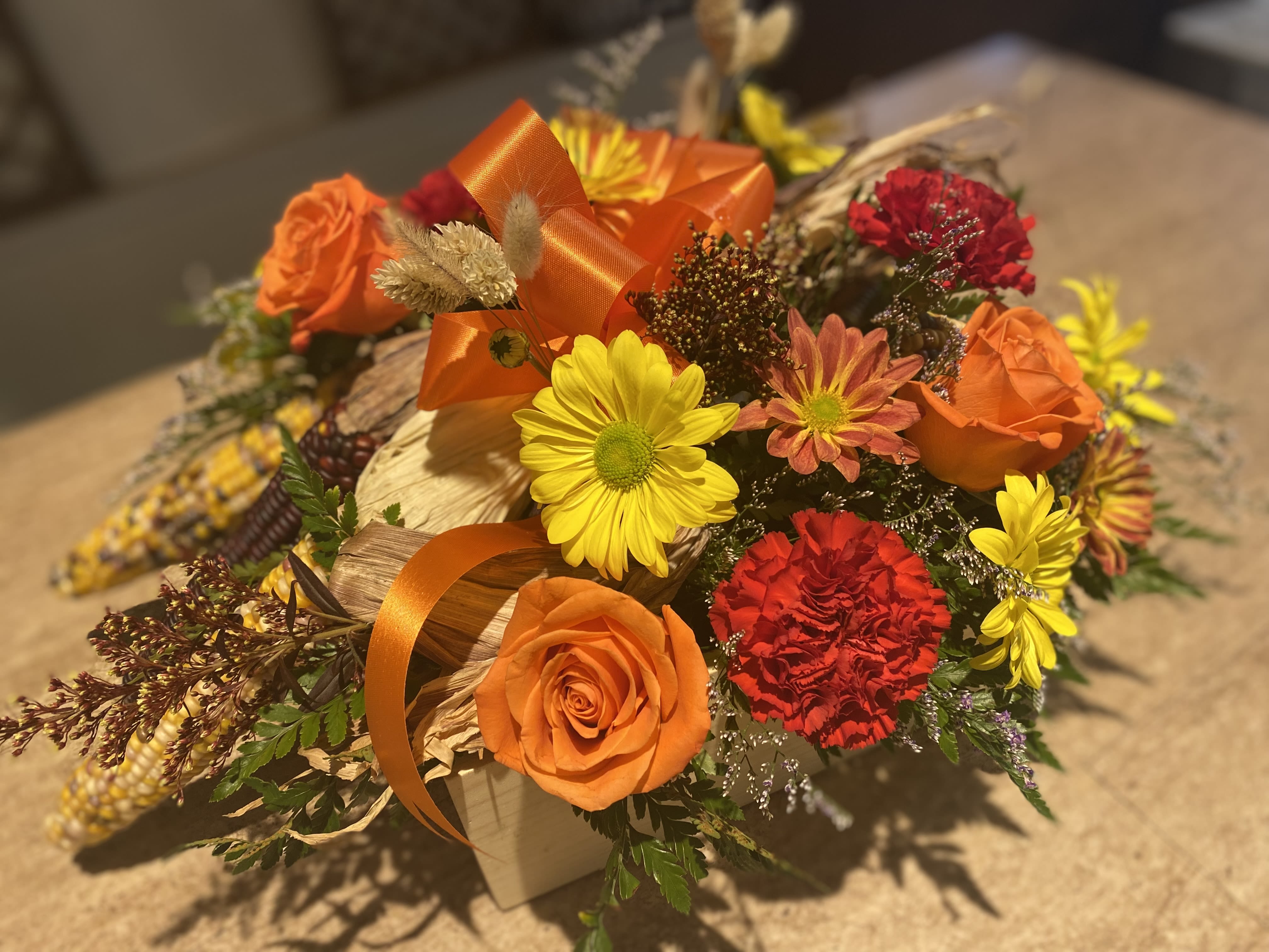 Indian Corn Autumn Flower Box Indian Corn Autumn Flower Box - A large wooden square box filled with Assorted Indian Corn, autumn pomps, hearty carnations, orange roses, fillers of solidago and limonium,  natural wheat and greenery topped with a fall colored bow in center.  Beautiful for the center of any table!