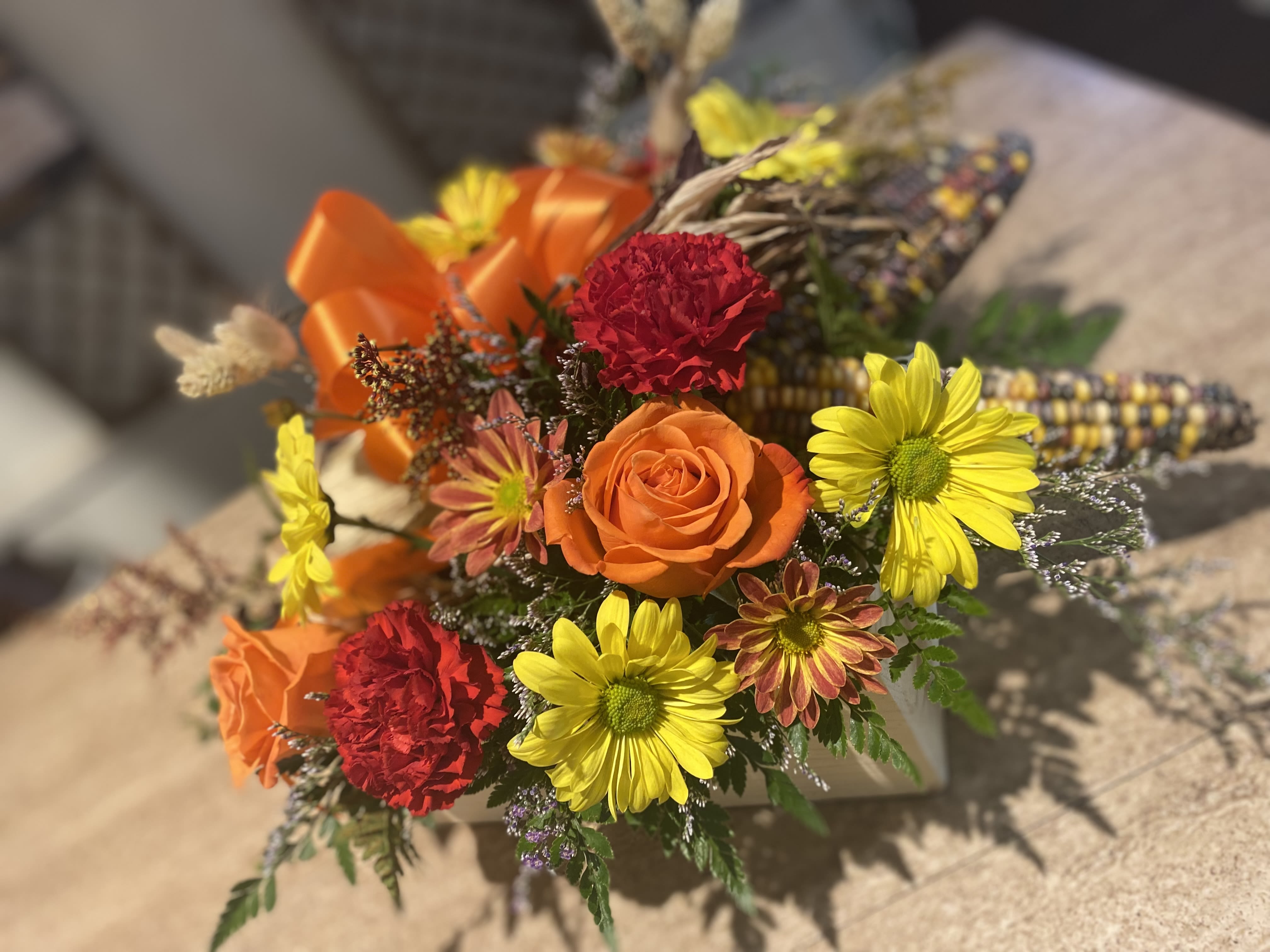 Indian Corn Wooden Flower Box Indian Corn Wooden Flower Box - A large square wooden box filled with assorted Indian Corn, hearty carnations, daisy mums, orange roses, solidago and limonium, greenery, natural wheat and a fall colored bow on top.  Beautiful for any table !!