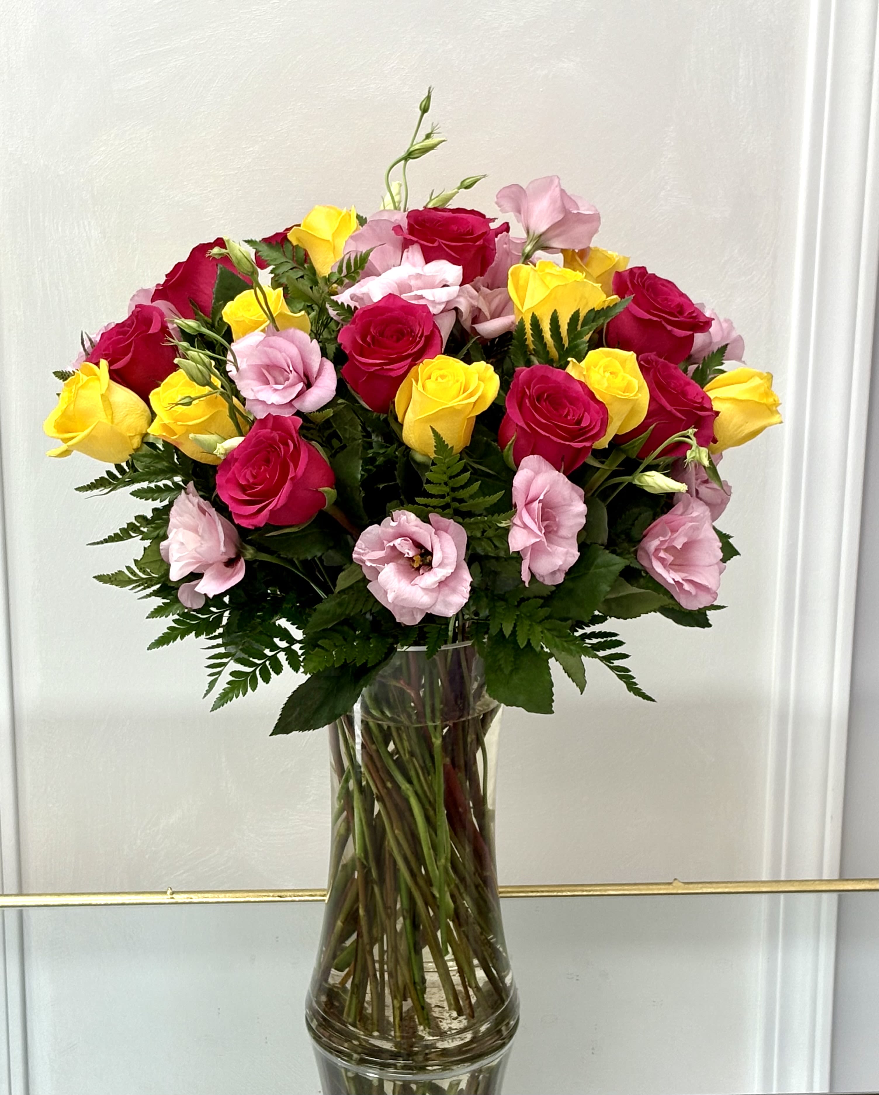 Sunshine Bloom By VLM Flowers - A joyful mix of pink lisianthus, hot pink roses, and yellow roses shines from a clear glass vase, blending warmth with elegance.