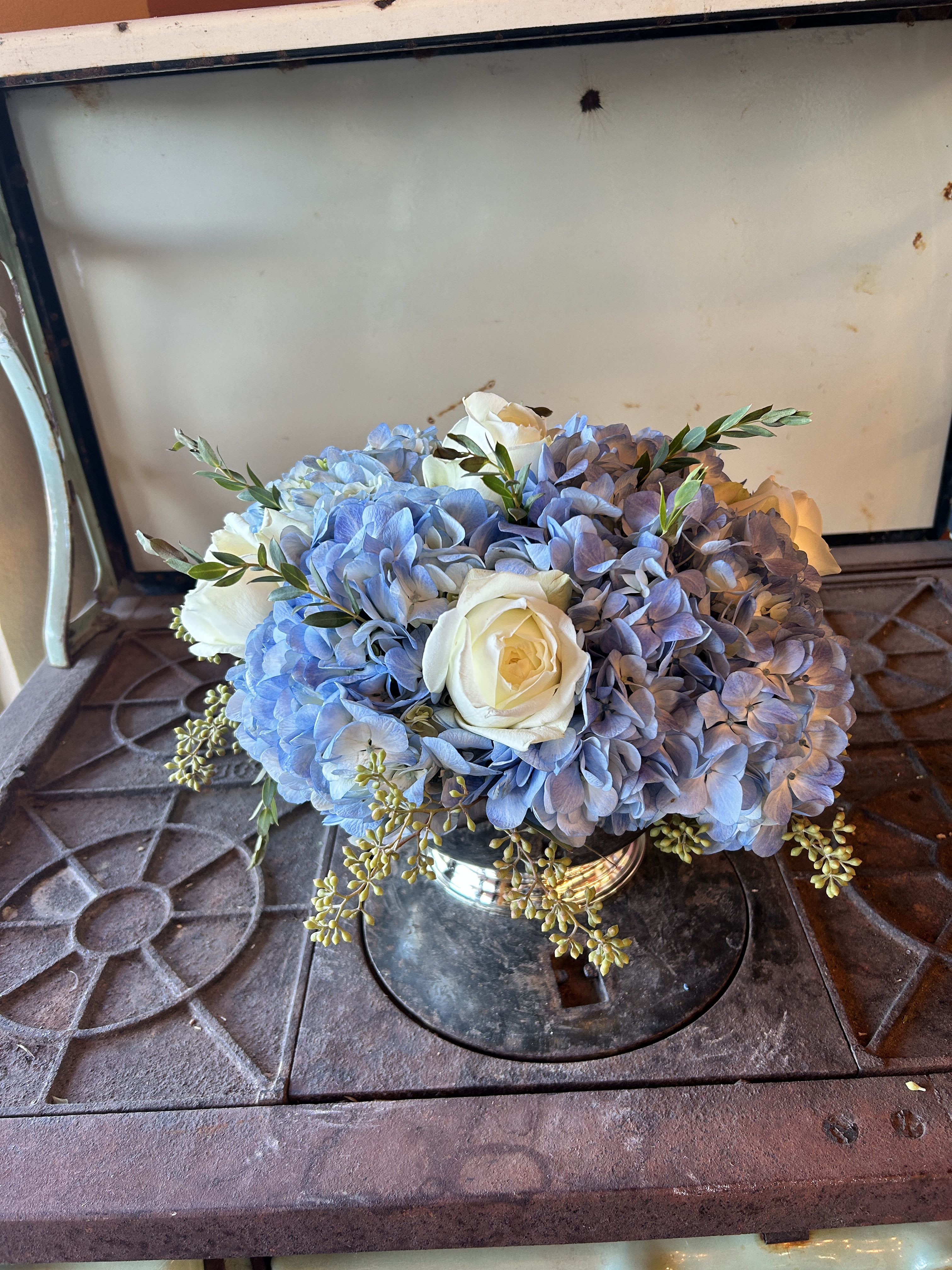 Blue Haven - Make any occasion unforgettable with this refined table arrangement featuring lush blue hydrangeas and graceful white roses, beautifully presented in a classic silver bowl. Accented with the soothing fragrance of fresh eucalyptus varieties, this design exudes tranquility and timeless sophistication. Perfect for weddings, dinner parties, or simply adding a touch of elegance to your home.