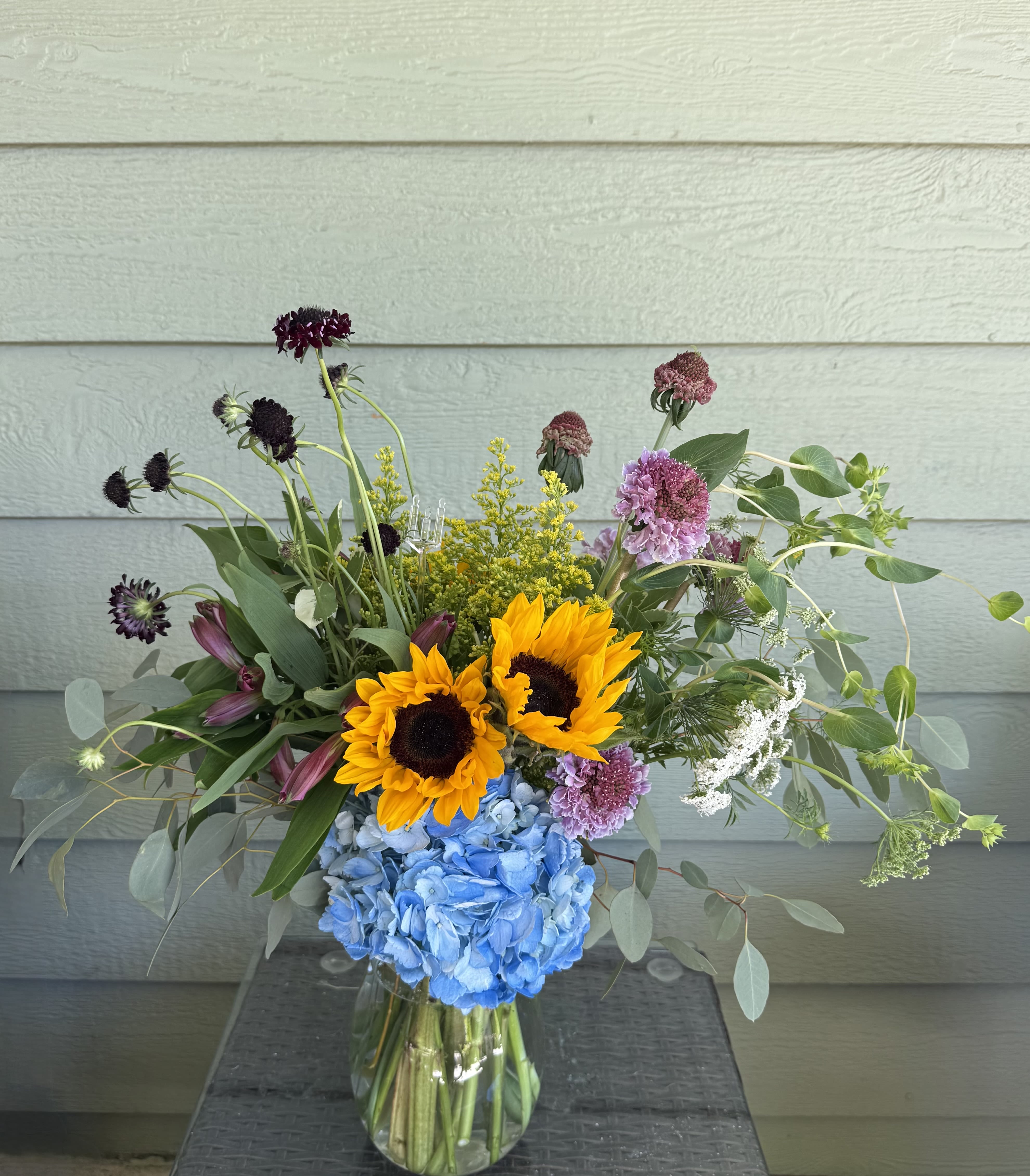 Modern Love - Celebrate the beauty of connection with Modern Love — a vibrant floral arrangement that redefines romance for today. Bold, joyful blooms like radiant sunflowers, sculptural scabiosa, and crisp blue hydrangea come together in an artful, asymmetric design, paired with lush greenery and unexpected textural accents. Housed in a sleek glass vase, this bouquet speaks the language of love with freshness, color, and confidence. Perfect for anniversaries, new beginnings, or just because — because modern love deserves modern flowers.