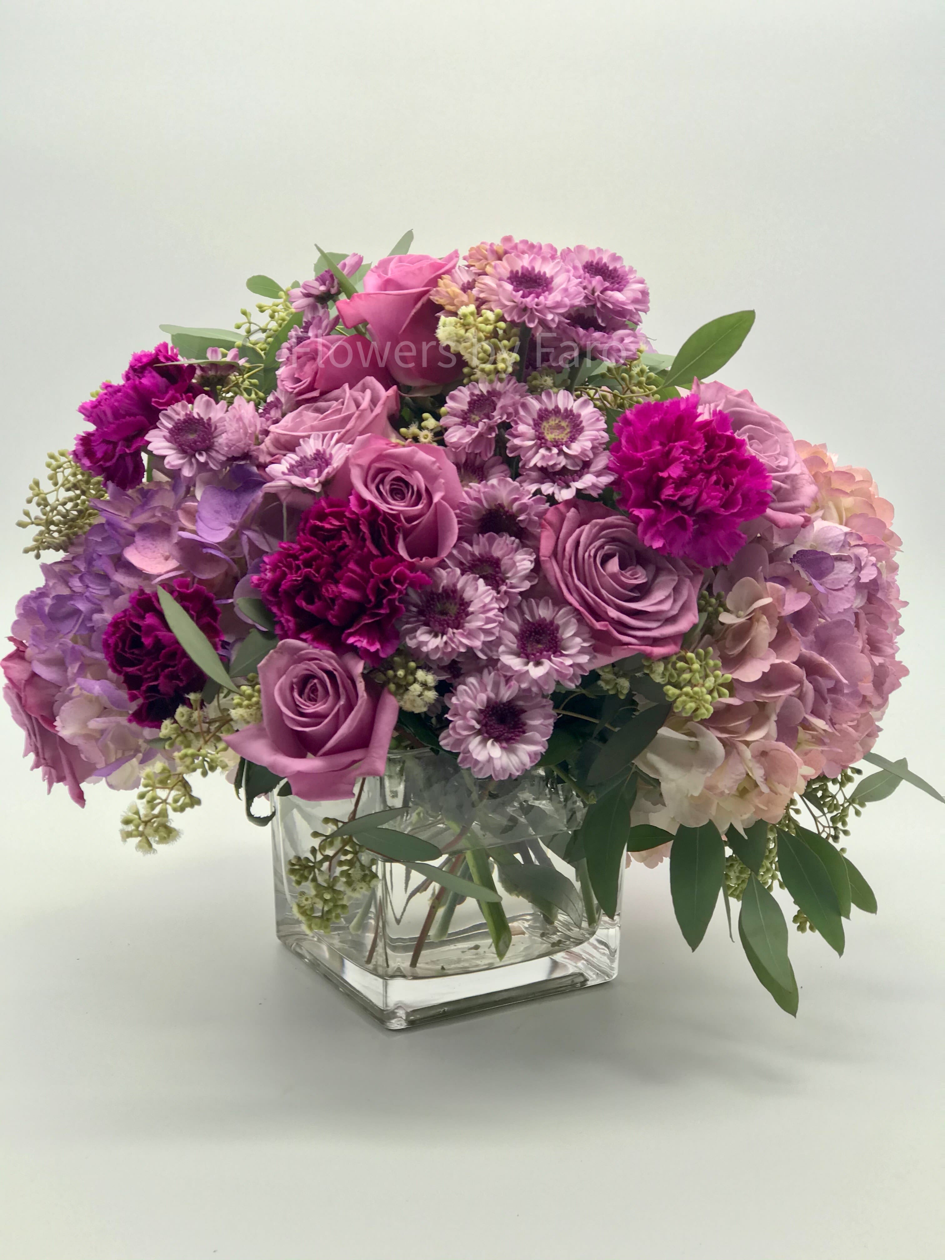 Lavender Joy - The Lavender Joy Bouquet is an arrangement of Lavender flowers like Roses, Hydrangeas, Carnations accented with Pom Poms and greeneries in a Glass Cube Vase.