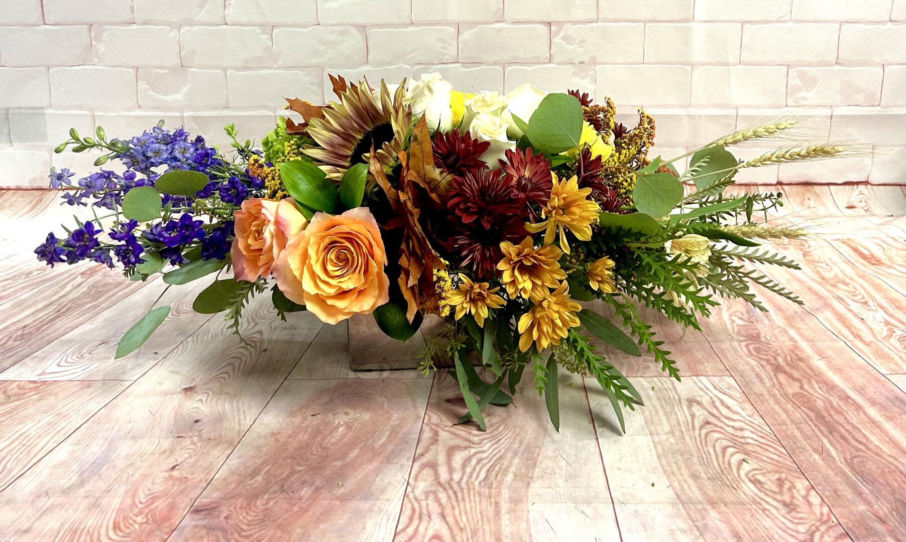 Golden Fields Thanksgiving Centerpiece - Inspired by the rich hues of harvest time, our Golden Fields Thanksgiving Centerpiece captures the warmth and abundance of the season. Vibrant orange roses, golden-yellow mums, and burgundy blooms are paired with rustic sunflowers, blue delphiniums, and natural wheat for a stunning mix of color and texture. Designed to bring the beauty of the autumn countryside to your table, this handcrafted arrangement is perfect for Thanksgiving gatherings or any fall celebration.